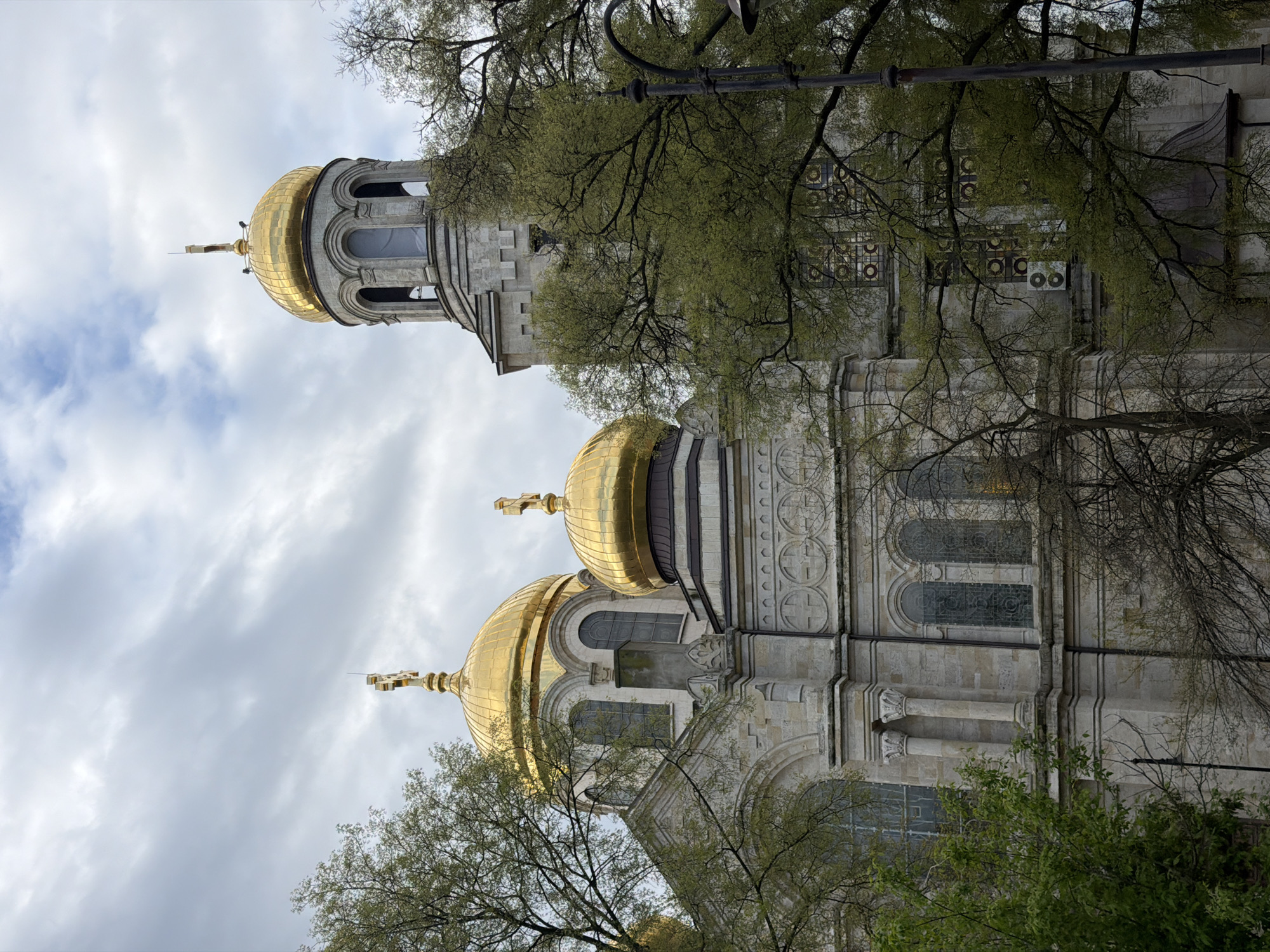 This image captures a grand Eastern Orthodox cathedral, identifiable by its multiple golden domes and intricate stone architecture. The main structure features a light-colored, possibly grey or beige, stone facade, adorned with decorative elements such as arched windows, elaborate moldings, and circular patterns.
Three prominent golden domes are visible, two of which are large and positioned in the foreground, each topped with a golden Orthodox cross. A taller bell tower rises behind them, also crowned with a smaller golden dome and a cross, extending higher than the surrounding elements.
The cathedral is partially framed and somewhat obscured by trees with green foliage, suggesting a season of spring or early summer. The branches, some bare and some covered in fresh leaves, intertwine around parts of the building. A black metal street lamp is partially visible in the upper right section of the image.
The sky is partly cloudy, with a mix of soft grey clouds and brighter areas, indicating a daytime scene, likely late morning or early afternoon. The lighting is diffused, illuminating the golden domes with a soft sheen. No people or specific activities are depicted in the scene, focusing solely on the architectural beauty and majesty of the building. The image's orientation is vertical. No text is visible. Given the location context, this is the Dormition of the Theotokos Cathedral in Varna, Bulgaria.