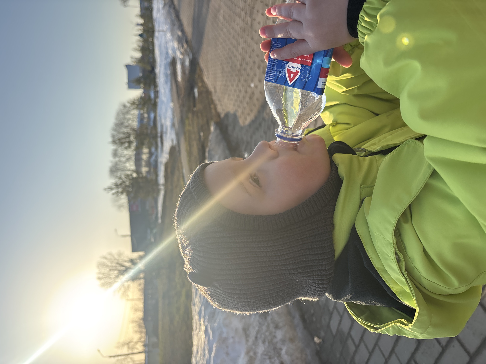 An outdoor scene in Chaikovsky, Russia, depicts a young child, seen from the chest up, drinking from a plastic bottle. The child is wearing a dark grey knitted balaclava-style hat with two small ear-like protrusions and a bright neon green jacket over a dark inner layer. The clear plastic bottle, held by the child's left hand, contains a clear liquid and features a blue cap and a blue and red label with the brand "AQUALITY" and Cyrillic text visible. The child's head is tilted back, and the bottle's opening is at their mouth. The immediate foreground shows a light-colored paved surface, possibly brick or concrete pavers. Beyond the child, the middle ground features patches of melting snow, bare deciduous trees, and evergreen trees on an unkempt ground. The background includes several blurred buildings, notably a darker, larger structure on the right, and two discernible streetlights. A strong, low sun is positioned in the upper left, casting significant light and lens flare across the scene, indicating either early morning or late afternoon.