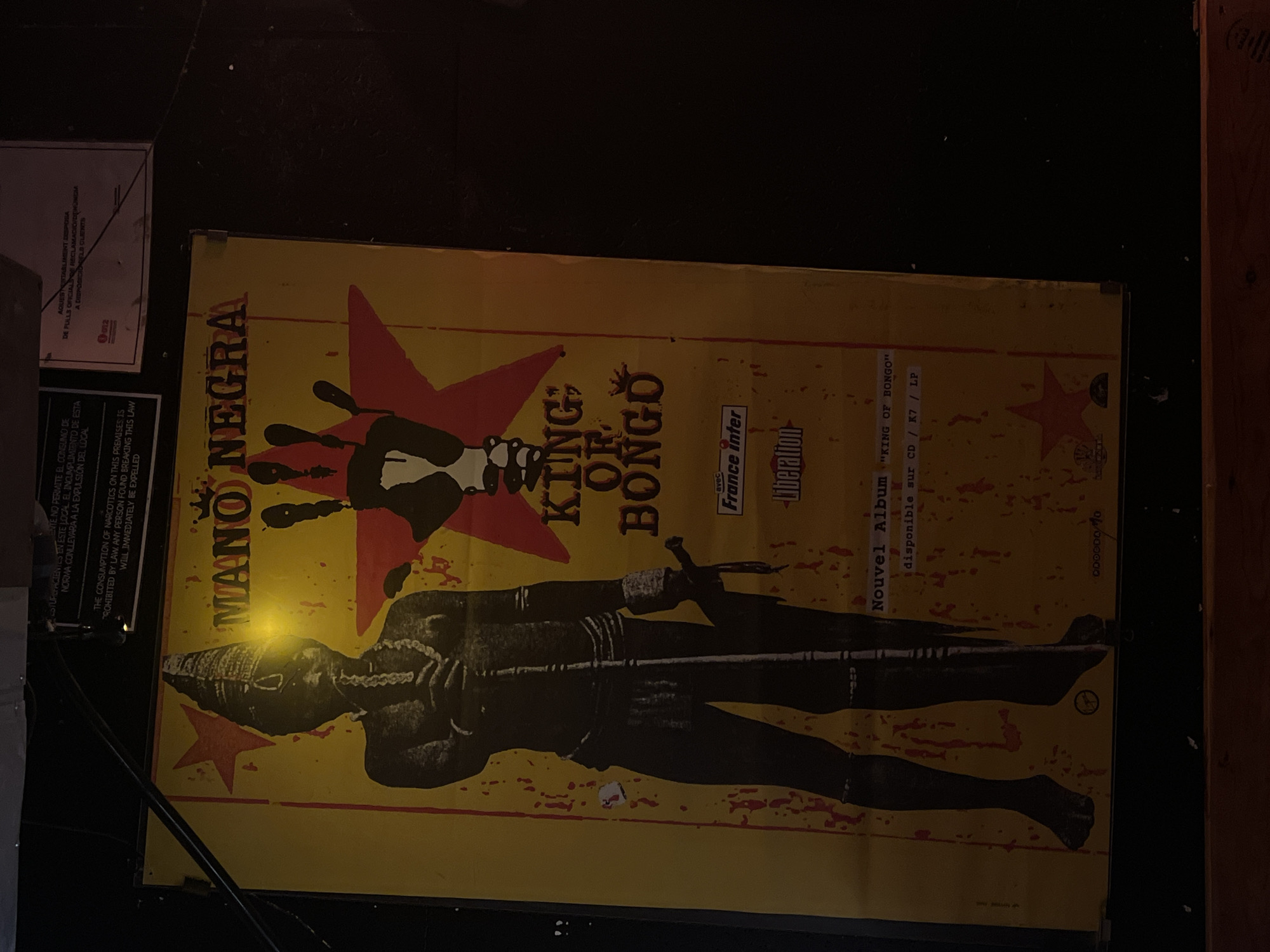 A poster for the band Mano Negra is displayed on a wall. The poster features a stylized image of a person wearing a tall headdress and holding a staff, with a large red star in the background. Text on the poster announces their album "King of Bongo". Small notices, possibly public health warnings, are affixed to the wall above the poster. The lighting suggests an indoor, possibly dimly lit environment. The poster is located in Barcelona, Spain.