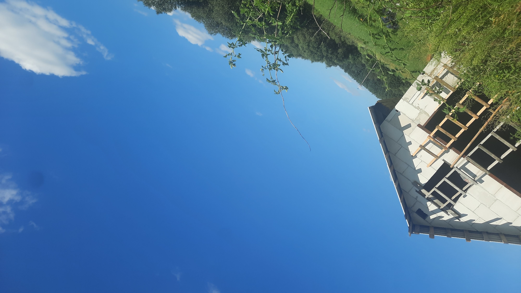 The image depicts an unfinished building, likely a house, constructed from concrete blocks, in a rural setting. The building is tilted at an angle, with wooden scaffolding visible against its exterior. The roof appears to be partially constructed. The surrounding environment is characterized by lush green grass and trees, suggesting a natural or semi-rural landscape. The sky is a vibrant blue with scattered white clouds. The presence of an unfinished structure implies ongoing construction or a project in progress. The location is identified as Shklou, Belarus.