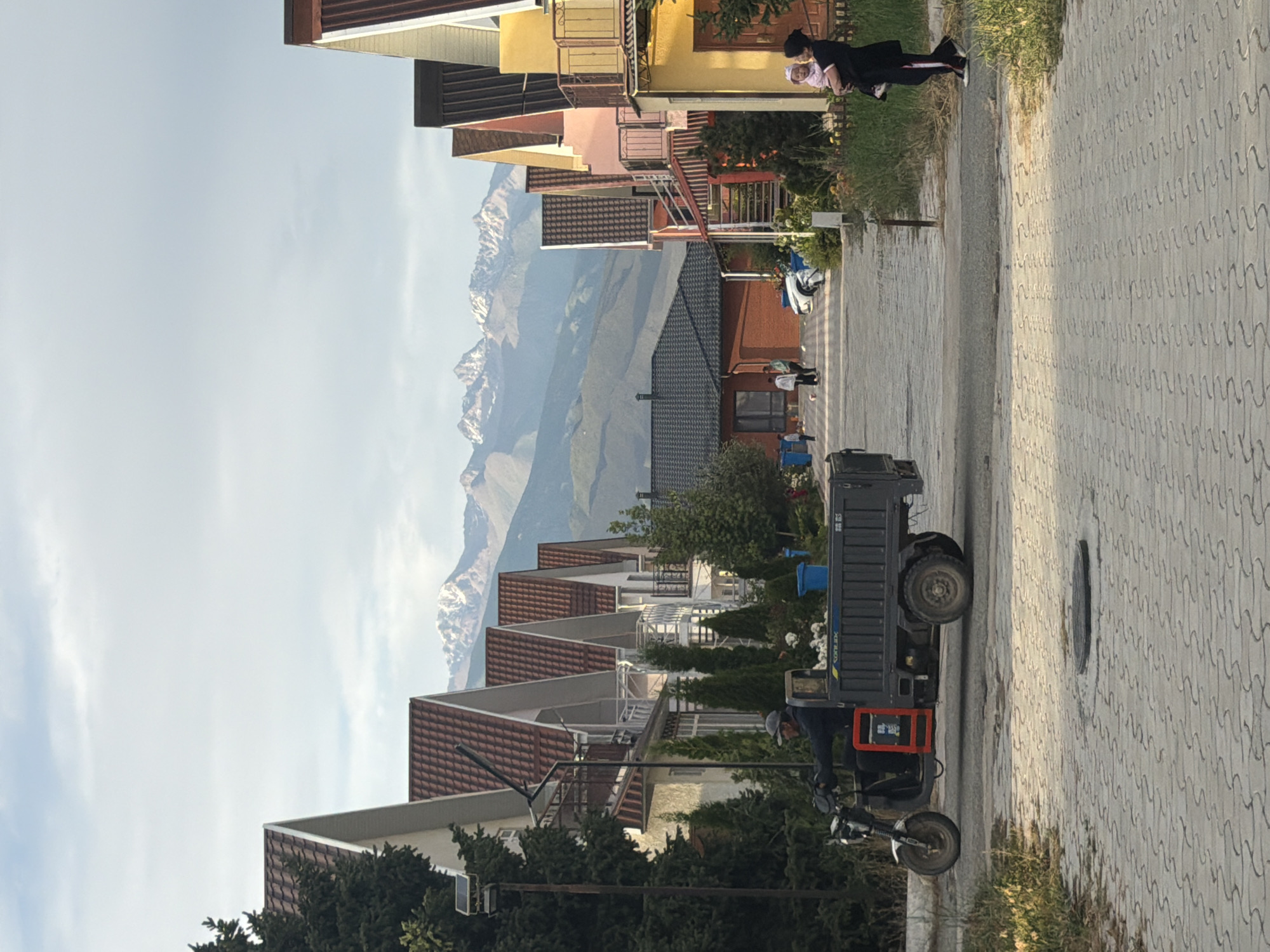 The image shows a street with houses and mountains in the background. A woman is holding a baby in her arms, standing on a sidewalk next to a row of houses. A small truck is parked on the street, and a person is sitting on a motorcycle in front of it. The houses are modern with pitched roofs and balconies. The mountains are snow-capped and appear to be quite large. The sky is partly cloudy, and the time of day appears to be late afternoon or early evening. The overall impression is one of a peaceful residential area with a beautiful natural backdrop. There is some faint text on the side of the truck, but it is not legible.