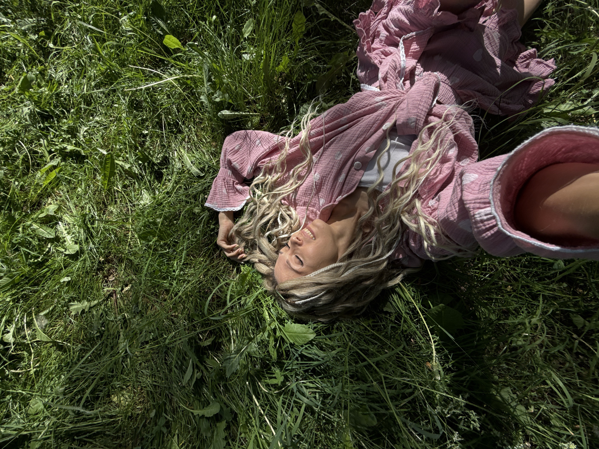 A high-angle, outdoor shot depicts an adult female lying supine on a dense patch of green grass in Rusanovo, Russia. The woman, positioned diagonally with her head towards the upper left, has fair skin, closed eyes, and a slight smile. Her long, wavy blonde hair contains several white or light-colored braided sections. She wears a pink, textured two-piece garment, possibly made of gauze, featuring a subtle white heart pattern. A white undergarment is visible beneath the buttoned top. The garment's sleeves and skirt feature ruffled edges with white lace trim. Her right hand is positioned near her head, fingers intertwined with her hair, while her left arm extends downward, with the hand partially visible at the bottom of the frame. The surrounding environment consists of tall, unkempt green grass with various broad-leafed weeds. Strong sunlight casts distinct shadows across the grass, illuminating the woman's face and parts of her clothing.