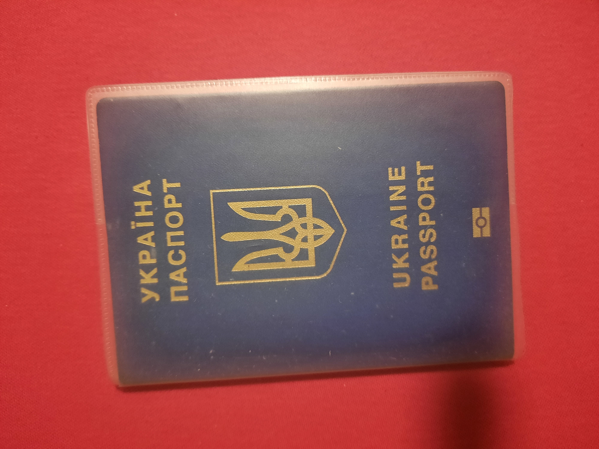 A dark blue Ukrainian passport, enclosed in a clear plastic cover, lies on a vibrant red surface. The passport displays the Ukrainian coat of arms in gold, a trident emblem, above the words "UKRAINE PASSPORT" also in gold lettering. The passport cover is slightly creased. The red surface appears to be fabric, possibly a tablecloth or a piece of clothing, and is illuminated from above, casting a subtle shadow to the left of the passport. The overall scene is static and presents a close-up view of the document. There is no indication of the city of Rosenheim, Germany, or any specific location within it.