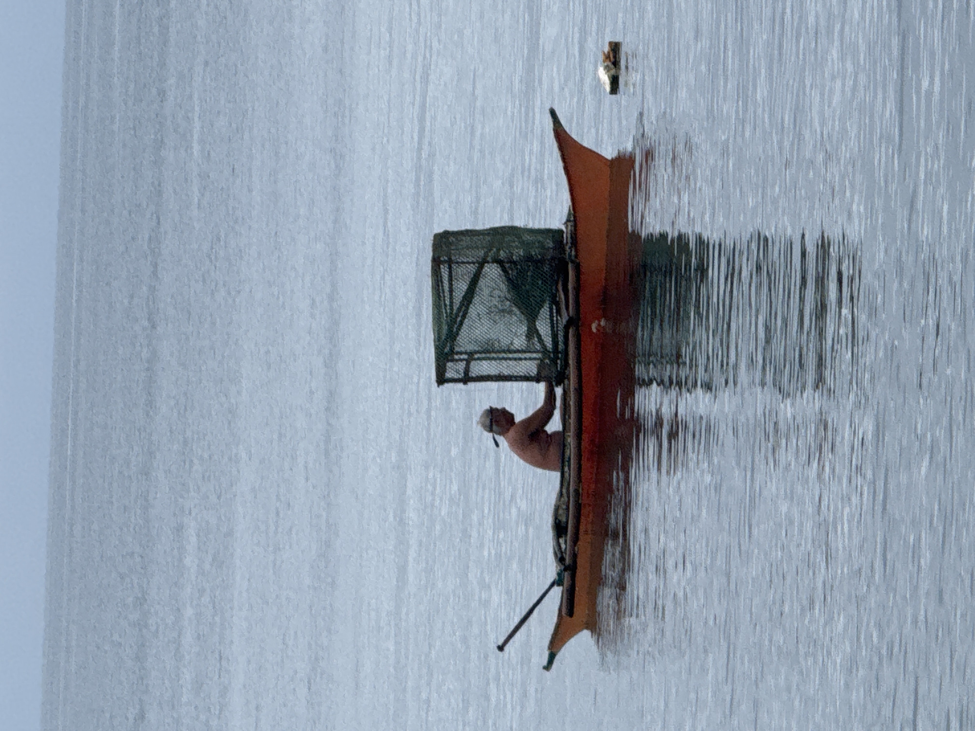 A man is standing in a narrow orange boat on the water. He is holding onto a square metal cage structure with a green mesh. The water appears calm with some rippling. The sky is overcast. The location is presumed to be Davao City, Philippines, although the image itself provides no way to confirm this fact.