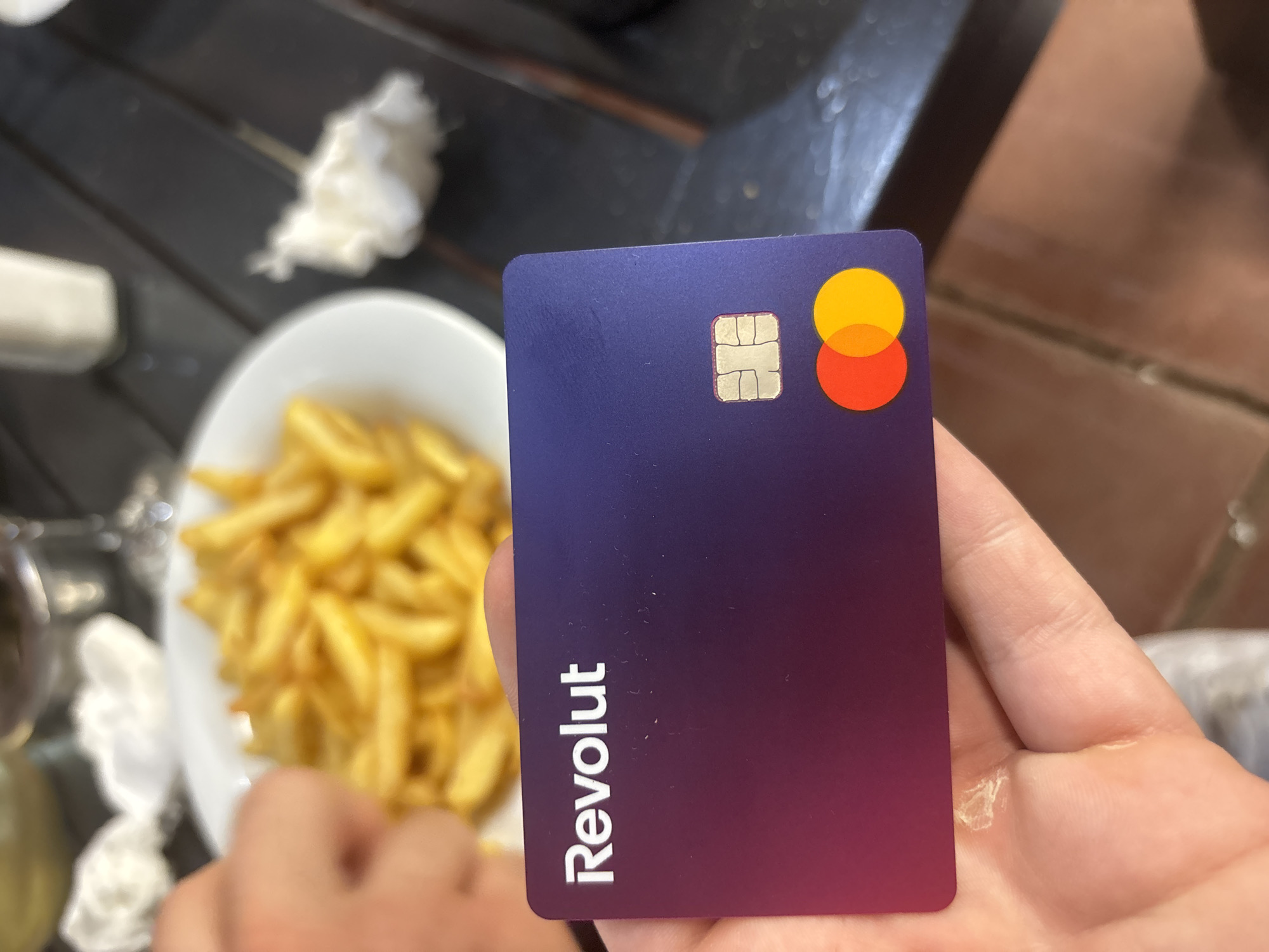 A close-up photograph captures a person's hand holding a Revolut Mastercard debit card, set within a restaurant environment in Blagoevgrad, Bulgaria. The card, held horizontally, features a gradient from dark purple at the top to a magenta-pink hue at the bottom. The "Revolut" logo is prominently embossed in white on the top left, with a gold-colored EMV chip located below it on the left side. The red and yellow interlocking circles of the Mastercard logo are visible on the bottom right corner of the card. The hand holding the card shows a small patch of dry or flaking skin on the palm near the base of the thumb. In the blurred background, a white plate filled with golden-yellow french fries occupies the center. Another hand, partially visible on the left, appears to be near the plate of fries. The items rest on a dark, slatted wooden table. To the right of the fries, a crumpled white napkin or tissue is visible on the table. Further in the background, out of focus, a clear glass (possibly containing a dark liquid) and a white shaker are discernible, alongside hints of a tiled floor or wall on the far right. The scene is illuminated by ambient light, suggesting an indoor or shaded outdoor dining setting.
