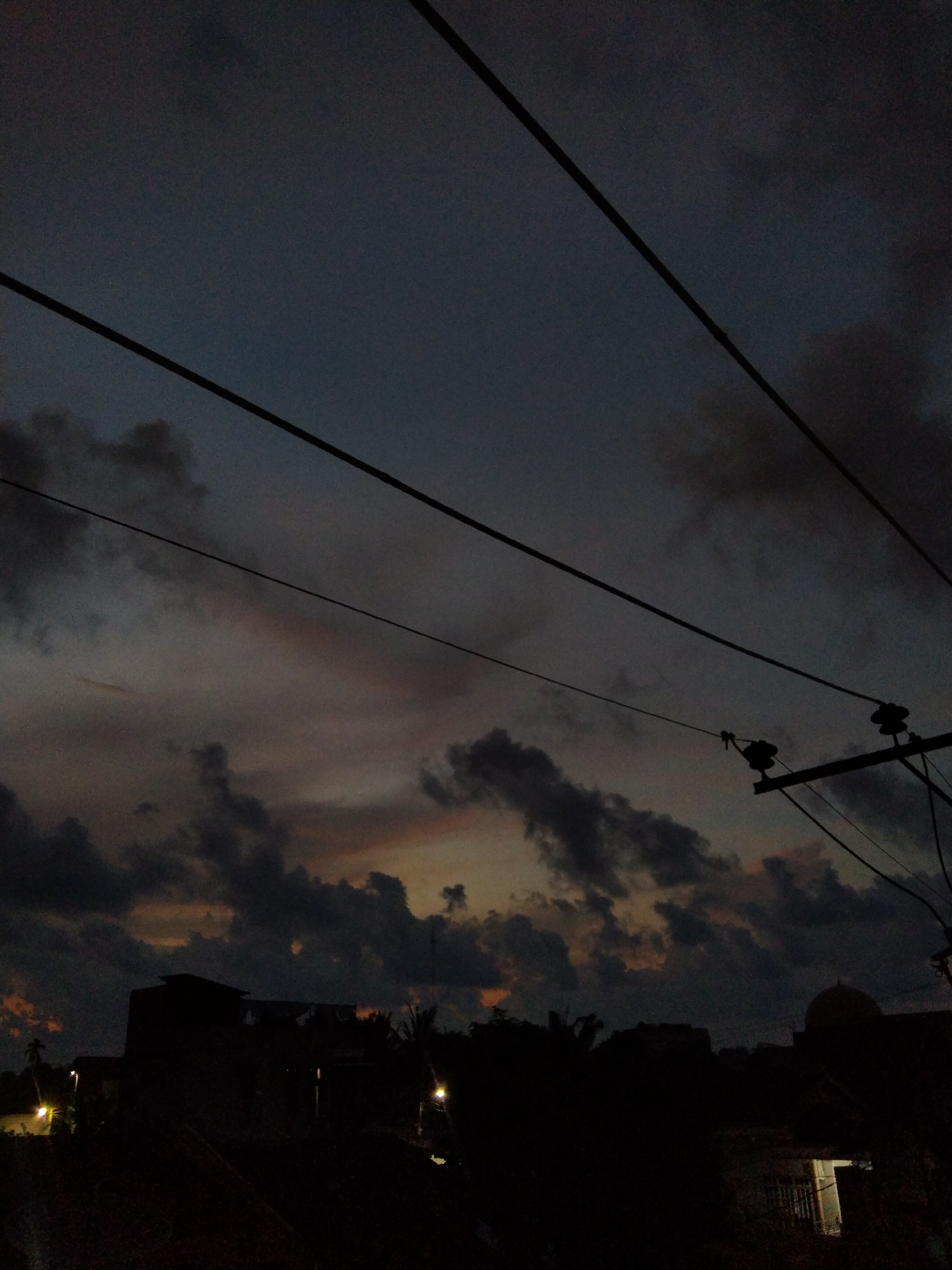 The image depicts a dusky scene in Bireuenmeunasahdayah, Indonesia. Overhead, electrical wires stretch across a sky filled with dark, clouded formations tinged with hints of orange light. Below, silhouetted buildings and foliage create a stark contrast against the fading light. A small, domed structure is visible amongst the buildings. A few scattered lights glow weakly in the darkness. The overall impression is one of impending nightfall in a quiet urban environment.