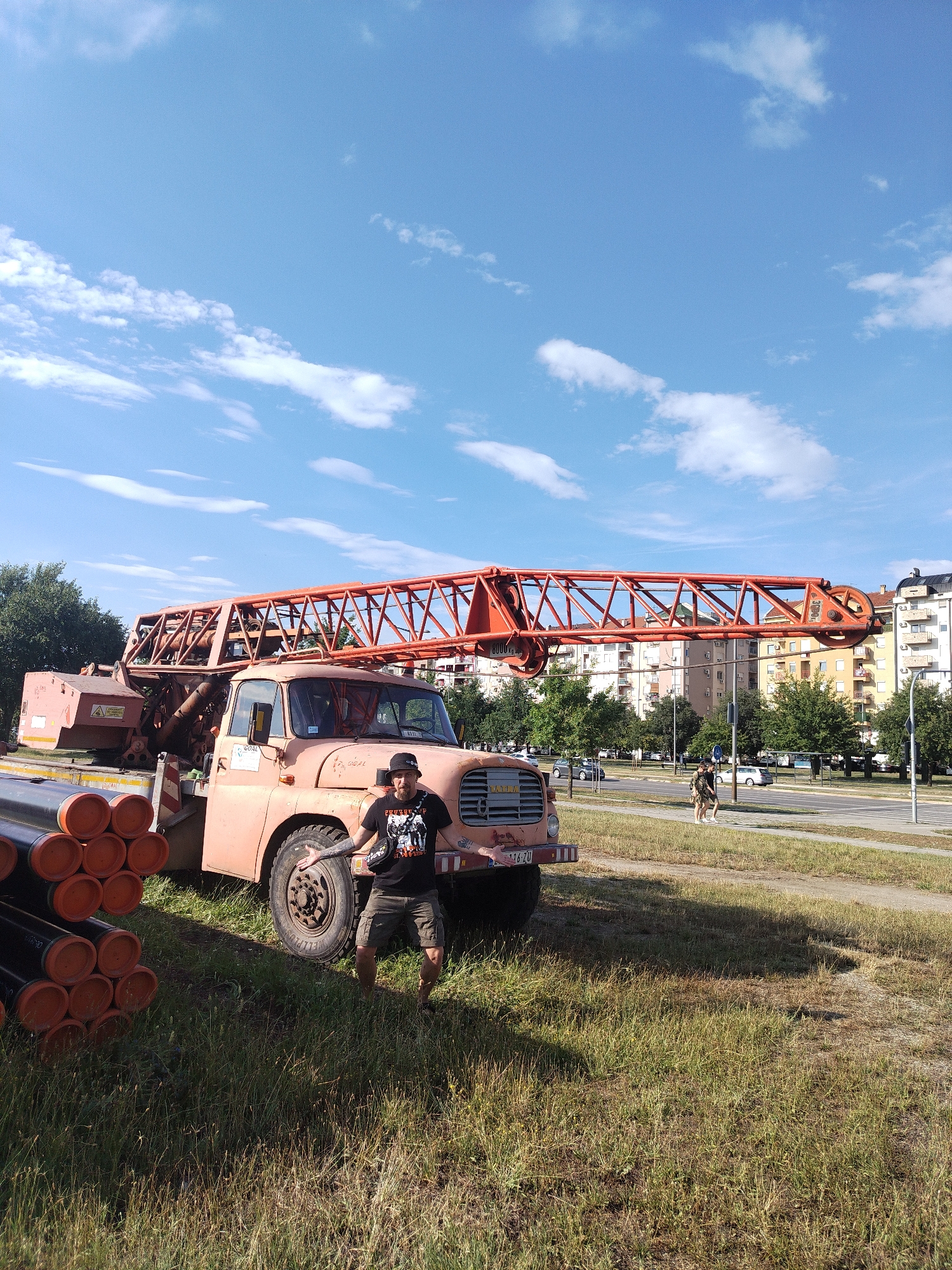 A man stands in front of a large orange crane truck parked on a grassy area in Novi Sad, Serbia. The truck is loaded with large pipes, stacked vertically to the left of the vehicle. The man is wearing a black t-shirt with a graphic print, camouflage shorts, and a black bucket hat. He is gesturing with his hands, palms open, towards the viewer. Behind the truck, buildings and trees are visible, suggesting an urban environment. The sky is blue with scattered white clouds. In the background, there is a road with a few cars and people walking. The scene appears to be outdoors on a sunny day.