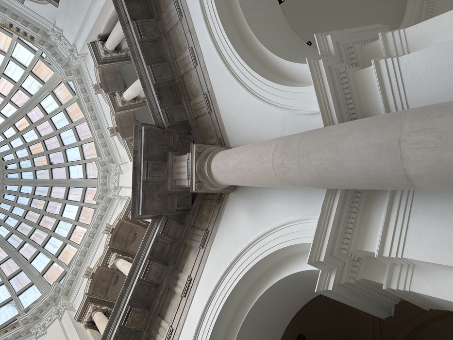 The image depicts the interior of a grand architectural structure, observed from a low angle looking upwards. The dominant feature is a multi-paned glass dome forming the ceiling, composed of numerous rectangular and square panels arranged in concentric rings, allowing natural light to illuminate the space. Below the dome, classical architectural elements are prominent, including a large, fluted, light-grey column in the foreground extending from the lower right towards the upper center. This column supports a complex entablature featuring decorative cornices and friezes. Further up, a series of smaller, similar columns with capitals are visible on an upper level, supporting another section of ornate stonework. Arches in a stark white finish are present at a lower level on the left, indicating a series of passageways or a vast open hall. The overall color palette is predominantly white and light grey, with subtle shadowing defining the architectural relief. No individuals, objects, or actions are depicted. This architectural style and specific dome structure are characteristic of the **Royal Exchange** building in the City of London, United Kingdom.