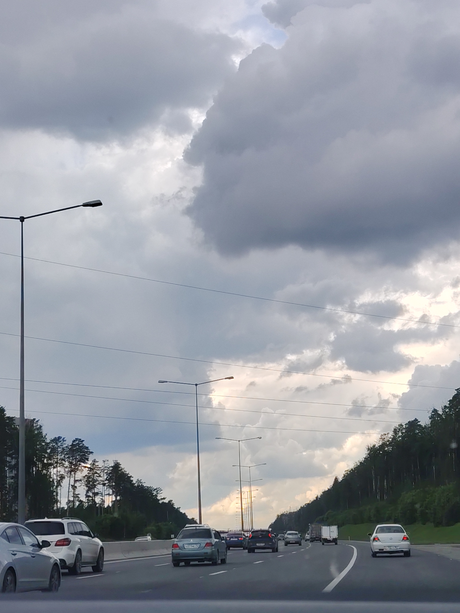 The image shows a highway with several cars driving on it under a cloudy sky. The road has multiple lanes and a concrete barrier separating traffic. On the left side of the road, there are tall streetlights and trees. On the right side of the road, there are more trees and a grassy embankment. The cars visible include a white SUV, a silver sedan, a light blue sedan, and a black hatchback. The license plate of the light blue sedan appears to be C371HC 66. The sky is filled with dark gray and white clouds. This scene is likely on the Ekaterinburg Ring Road (also known as the "Koltsevaya doroga" or E-22 highway) near Yekaterinburg, Russia, given the presence of Russian license plates and the typical infrastructure seen on Russian highways.