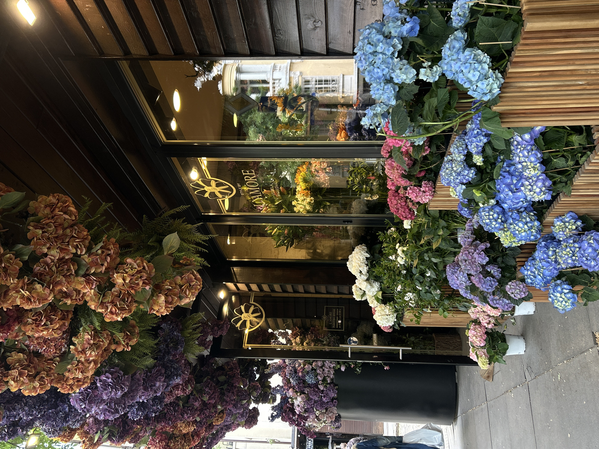 The image shows the exterior of L’AMORE, a flower shop in Sofia, Bulgaria. The shopfront is adorned with an abundance of hydrangeas in a variety of colors including blue, purple, white, pink, and brown. The flowers are arranged in planters. The shop's signage is visible on the glass. The reflection in the glass reveals a building across the street. The pavement is visible in the foreground. No people are clearly visible.