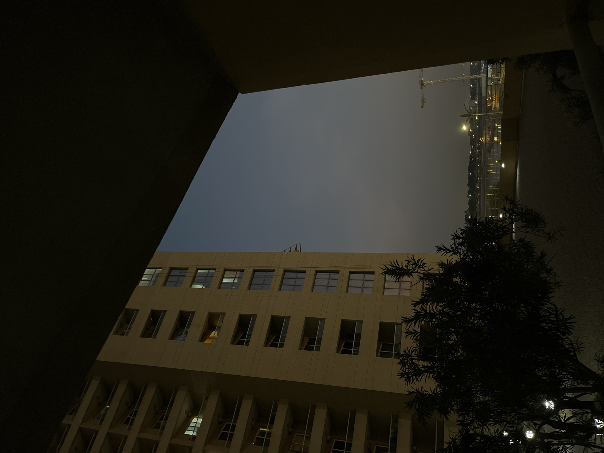 A nighttime view shows a tall, light yellow apartment building with balconies. The building is seen from a slightly low angle, with a dark overhang in the foreground. Across a body of water, a cityscape is visible with lights and a construction crane. The sky is a dusky, muted blue. In the foreground, a dark, leafy tree with small, illuminated lights is present. The overall impression is of an urban environment at twilight or after dark.