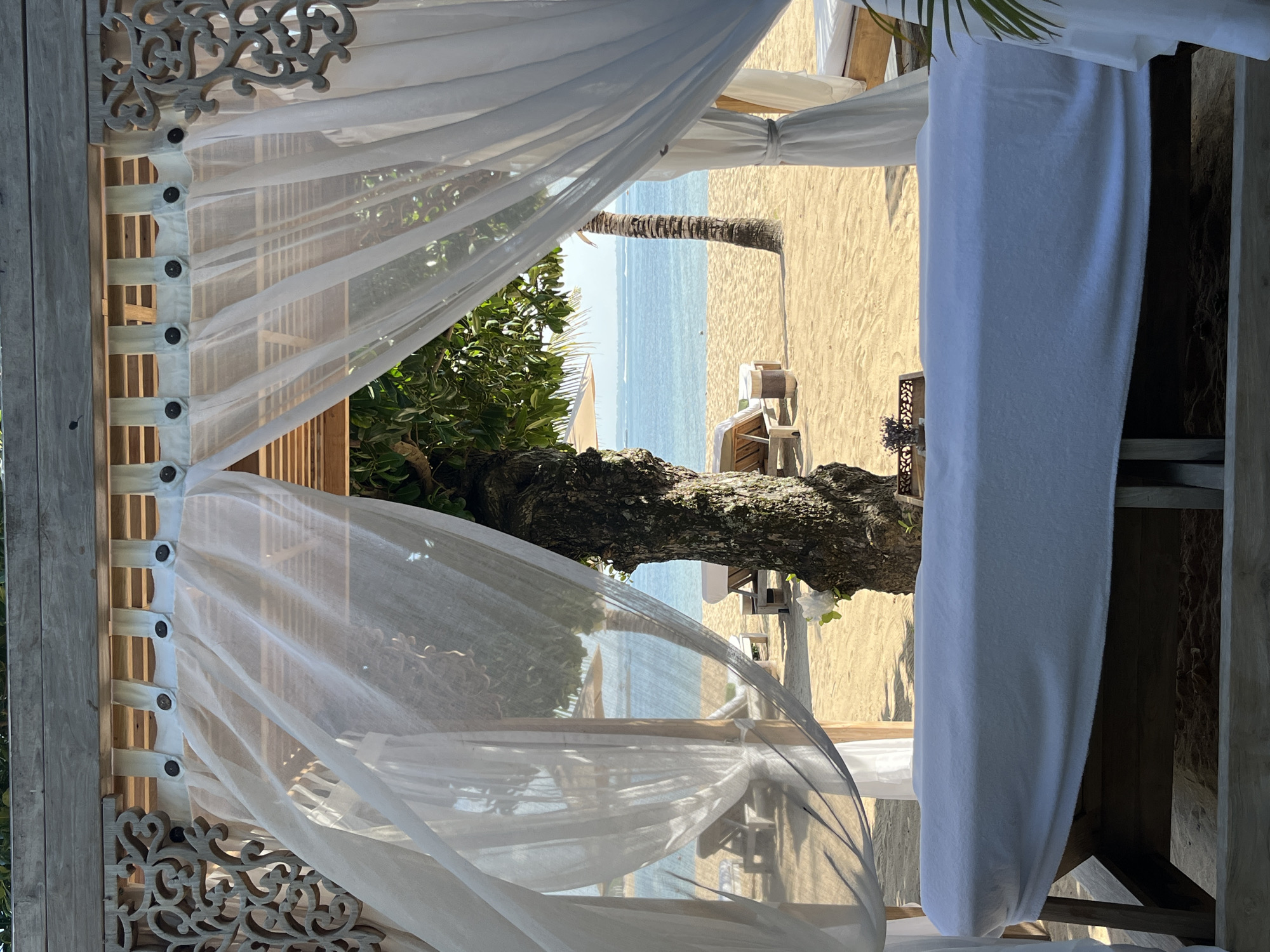 The image depicts a spa treatment area on a sandy beach in Kuta Selatan, Indonesia, overlooking the ocean. In the foreground, a massage table draped with a white sheet is visible under a wooden gazebo with white sheer curtains. Behind the table, a large tree trunk stands prominently, with green foliage above. The beach itself is dotted with lounge chairs and umbrellas, and the turquoise ocean stretches to the horizon under a clear sky. The overall scene evokes a serene and tranquil atmosphere, indicative of a luxury resort or spa setting.