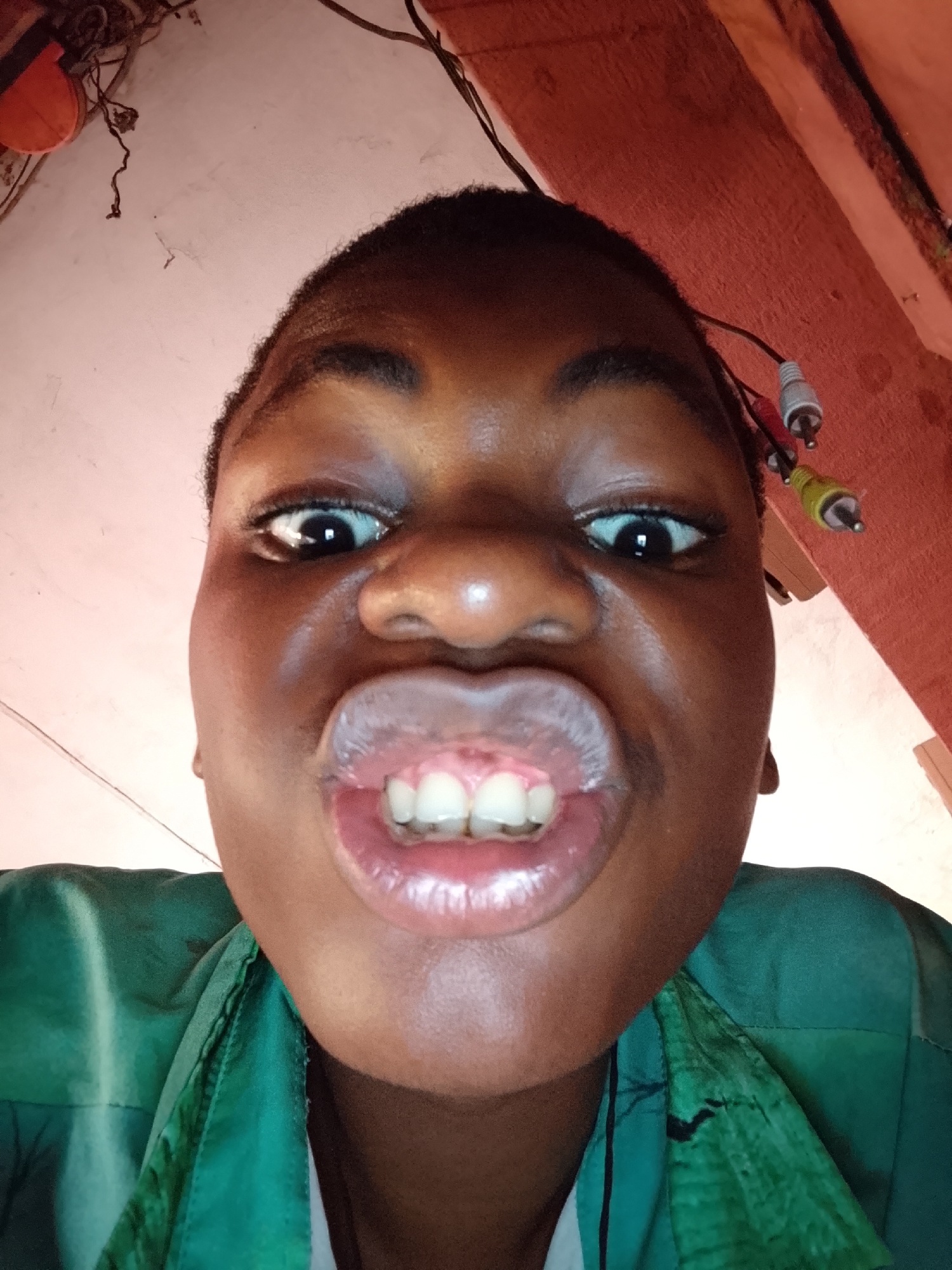 A close-up, low-angle shot features a person with dark skin making a playful facial expression. The individual is positioned centrally, looking directly at the camera. Their eyes are wide open, and their lips are puckered outwards, revealing their upper front teeth. Short, dark hair frames their face.
The person is wearing a dark green garment, possibly a shirt or jacket, visible across the lower portion of the frame. The background appears to be an indoor setting with a pale pinkish-beige wall or ceiling. Various wires and cables are visible, suggesting an exposed electrical setup. In the upper right, a cluster of RCA audio/video connectors (red, white, and yellow) dangles from a wire. To the upper left, an orange, somewhat circular object is attached to the wall, with more tangled wires around it. The overall impression is an informal, spontaneous moment. No text is visible.