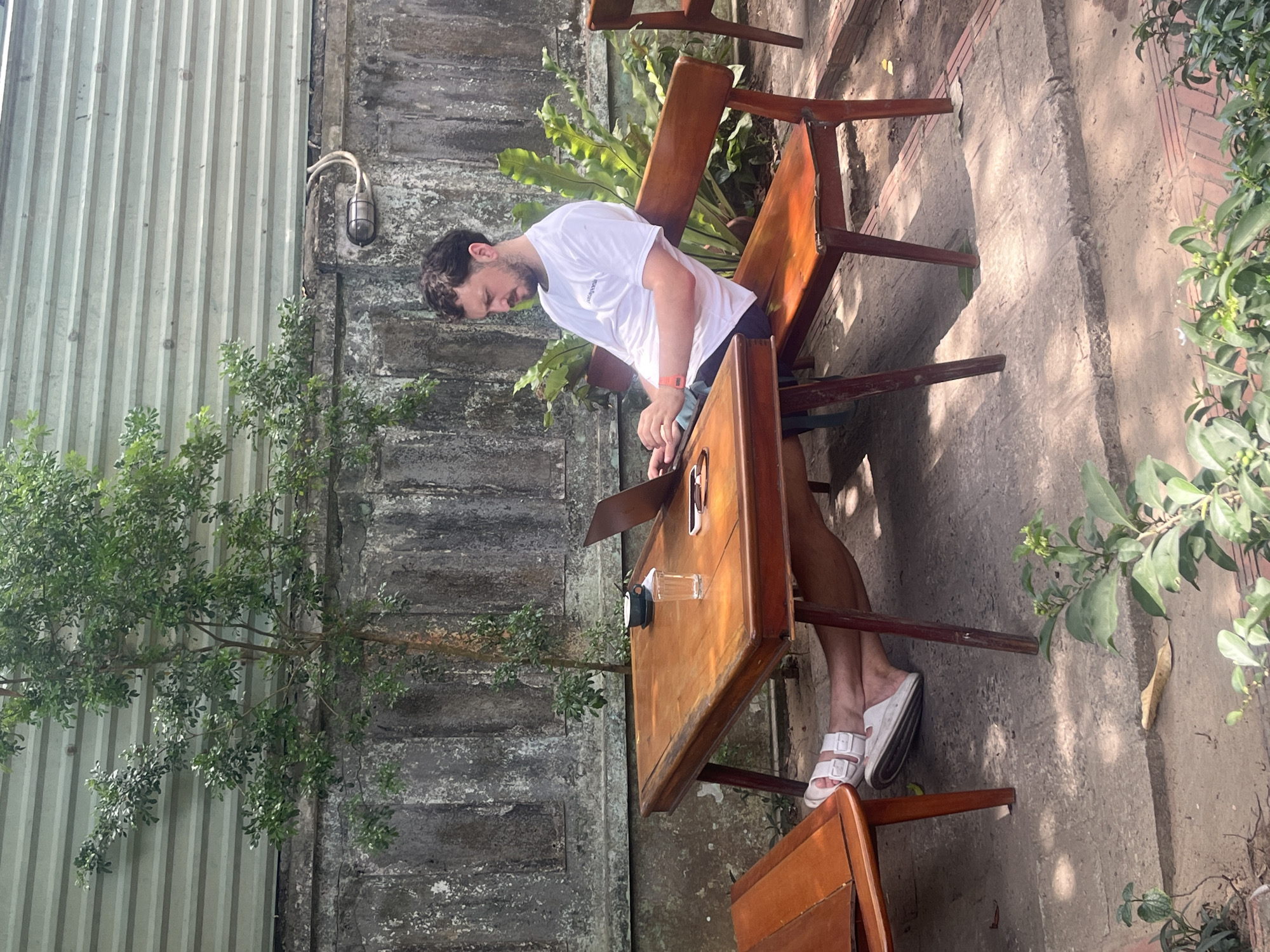 A man is sitting at a wooden table outdoors in what appears to be a cafe or outdoor seating area in Da Nang, Vietnam. He is wearing a white t-shirt and dark shorts with white slip-on sandals. The man is looking down at a laptop that is open on the table in front of him. To his left, there is a cup of coffee and a glass of water. Behind the man, there is a wooden bench and lush green plants. The background consists of a textured concrete wall and a corrugated metal structure. The ground is covered in a mix of dirt and paving stones.
