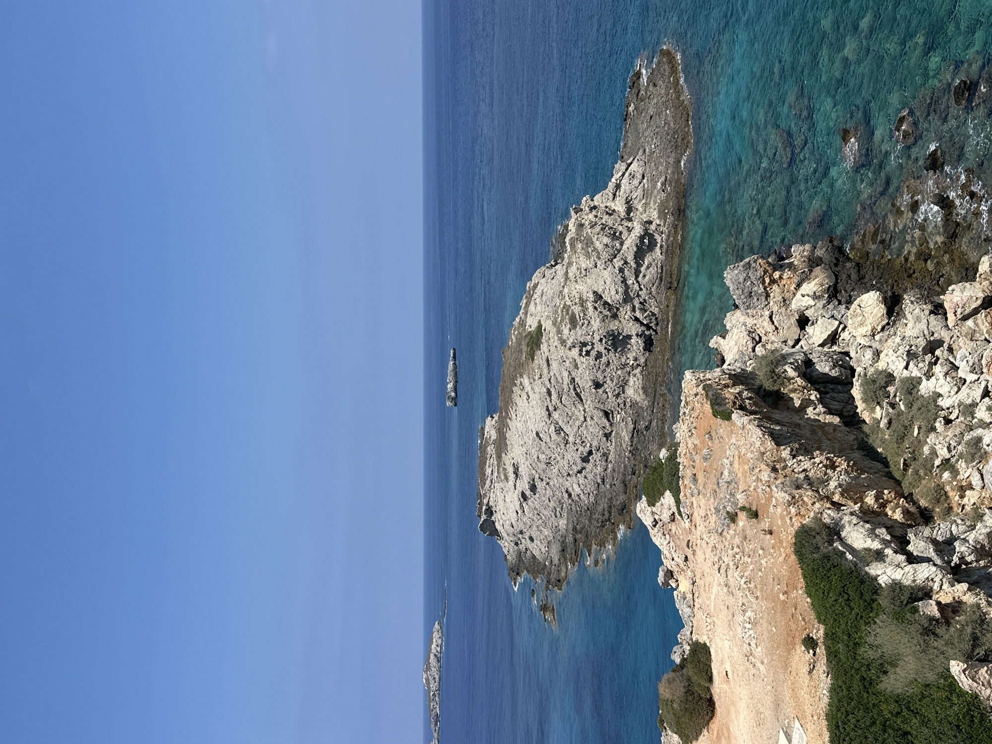 The image shows a scenic coastal landscape in Rizokarpaso, Northern Cyprus. The scene is dominated by a clear, bright blue sky and a vast expanse of deep blue ocean. In the foreground, a rocky coastline with rugged cliffs juts out into the sea. The rocks are varied in color, ranging from light gray to brown and are interspersed with patches of green vegetation, including bushes and shrubs. The water near the shore is a striking turquoise, revealing the rocky seabed beneath its surface.
Further out in the sea, several small, rocky islands are visible. The largest of these islands, situated in the mid-ground, is a prominent feature with a flat, rocky surface and sparse vegetation. In the distance, on the horizon, more islands can be seen, appearing as small, rugged shapes against the blue of the sea. A small, dark object, possibly a boat or a structure, is visible on the water in the distance. The overall impression is one of a remote and untouched natural environment. There are no people or animals visible in the image.