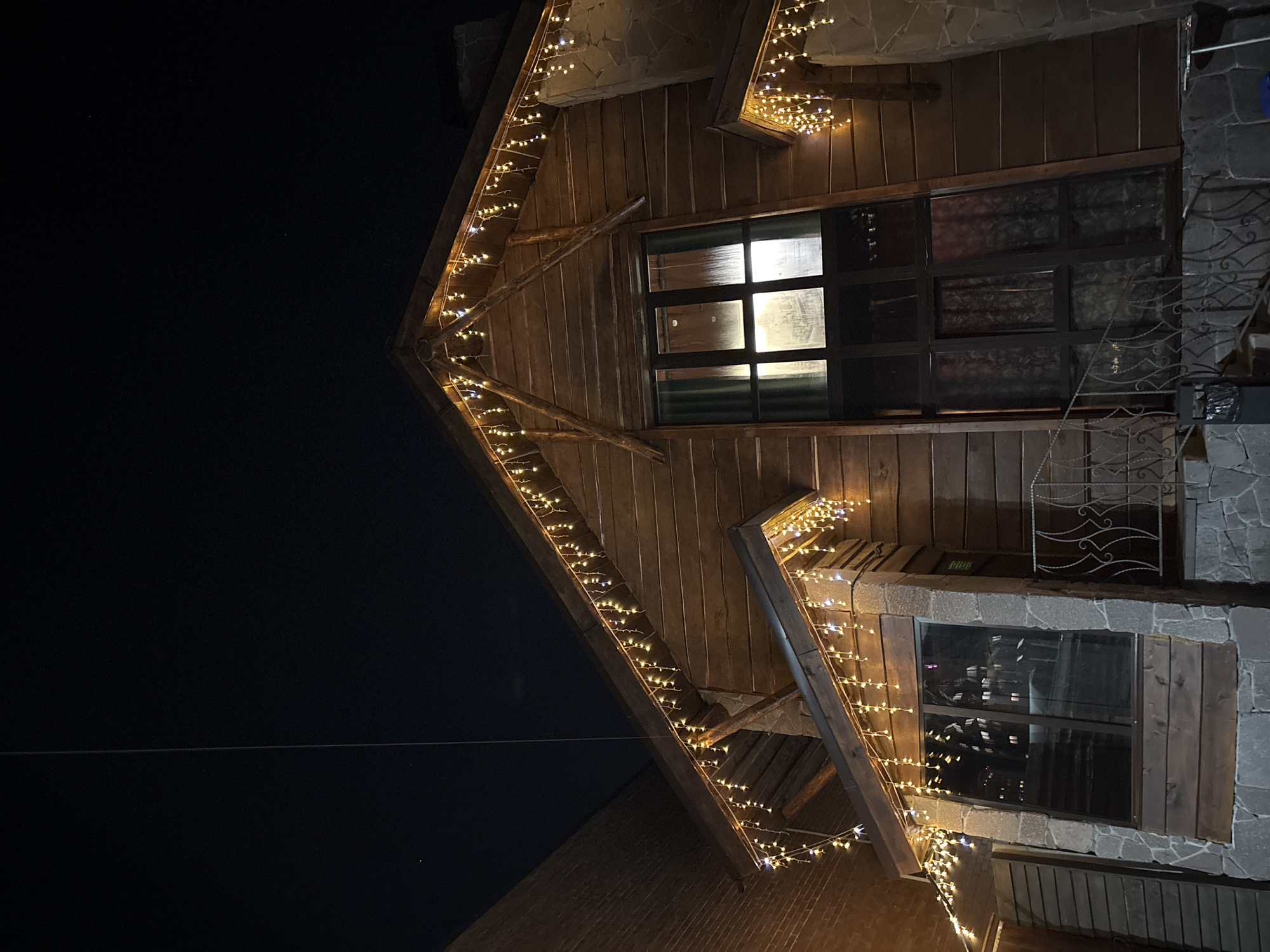 A low-angle, outdoor, nighttime photograph depicts a building's exterior. The building has dark-brown wood siding and is decorated with strings of small, warm-white lights along its roofline. The architectural style is angular, with a prominent peak.
The building features dark-framed windows. One window displays a grid pattern, and light is visible from inside. The building’s exterior also has stone elements. A small, decorative metal railing is visible near one window.
The background is dark, suggesting a night sky. A thin wire stretches across the dark area of the frame. The composition is shot from below, emphasizing the height and angles of the building.
There is no evidence in the image to place this image in Perm, Russia.