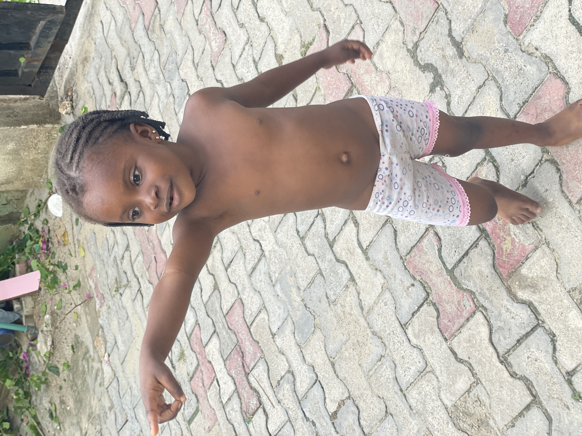 A young African girl stands on a patterned brick surface outdoors. She is wearing patterned shorts with pink trim. She has braided hair and small gold earrings. The background includes a partial view of a dark door and a patch of vegetation. The location is unknown.