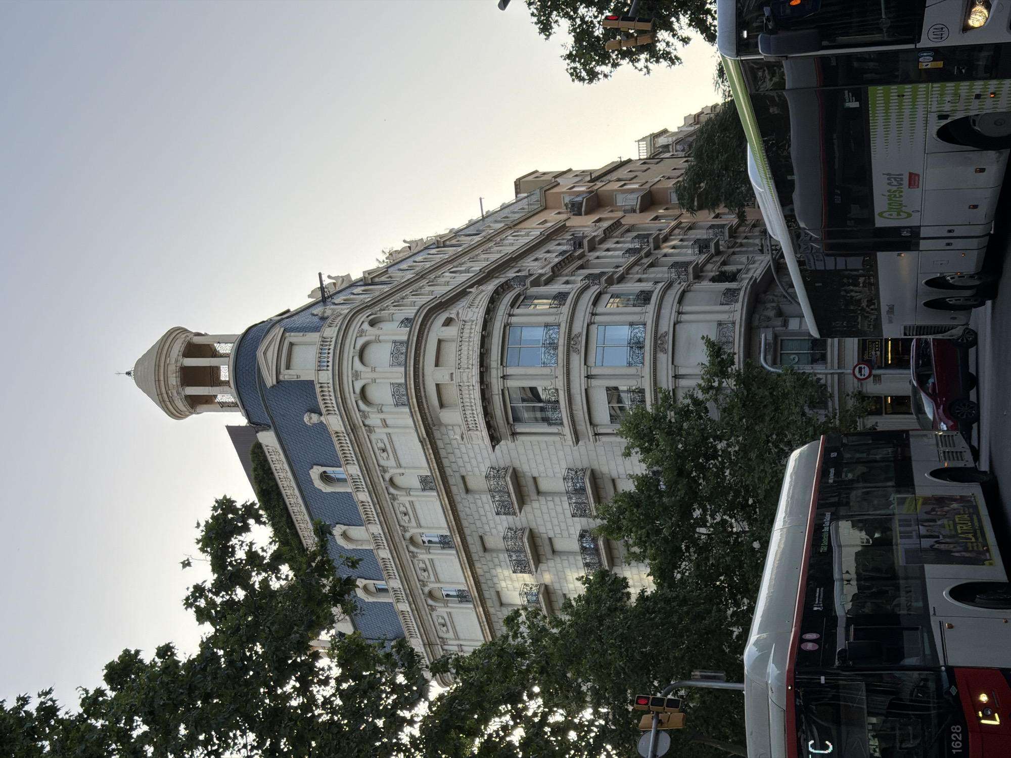 The image depicts a street scene in Barcelona, Spain, with a prominent, ornate building on the right and two buses in the foreground. The building features a mansard roof with dormer windows, a decorative cupola at the corner, and multiple balconies with intricate iron railings. The facade is constructed of light-colored stone blocks.
To the left, a red and white bus with the number "1628" is visible, displaying advertisements on its side. To the right, a white and green bus with "Exprès.cat" branding is also present. Both buses appear to be in motion. A red car is stopped behind the first bus.
Several trees with dense green foliage are scattered throughout the scene, obscuring parts of the building and the sky. Traffic lights are visible on a pole on the left side of the image. The sky is clear and pale, suggesting either early morning or late afternoon. The overall impression is of a busy urban environment with a mix of historic architecture and modern transportation.
