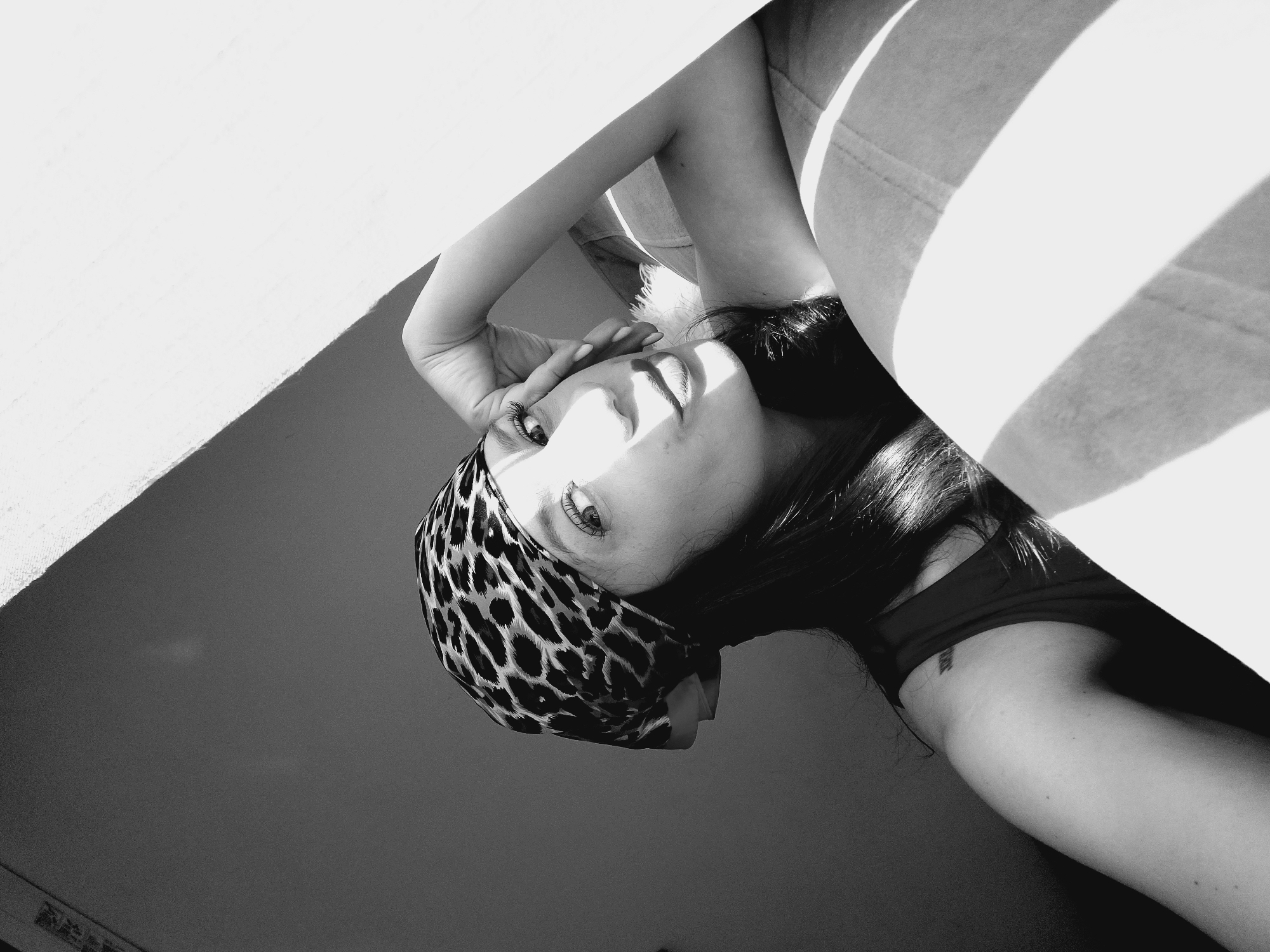 A black and white, medium shot photograph shows a young woman in Sofia, Bulgaria, lying on her side on a surface that appears to be a couch or bed. She is wearing a leopard print bandana on her head and a dark-colored tank top. Her right arm is propped up, supporting her head, with her hand resting on her forehead. The woman is looking at the camera with her lips slightly parted, and harsh sunlight is casting strong shadows across her face and the surrounding environment. The background is dimly lit, with a diagonal shadow from a window or light source creating a distinct line across the scene. The overall impression is one of a casual, perhaps intimate, moment captured in natural light.