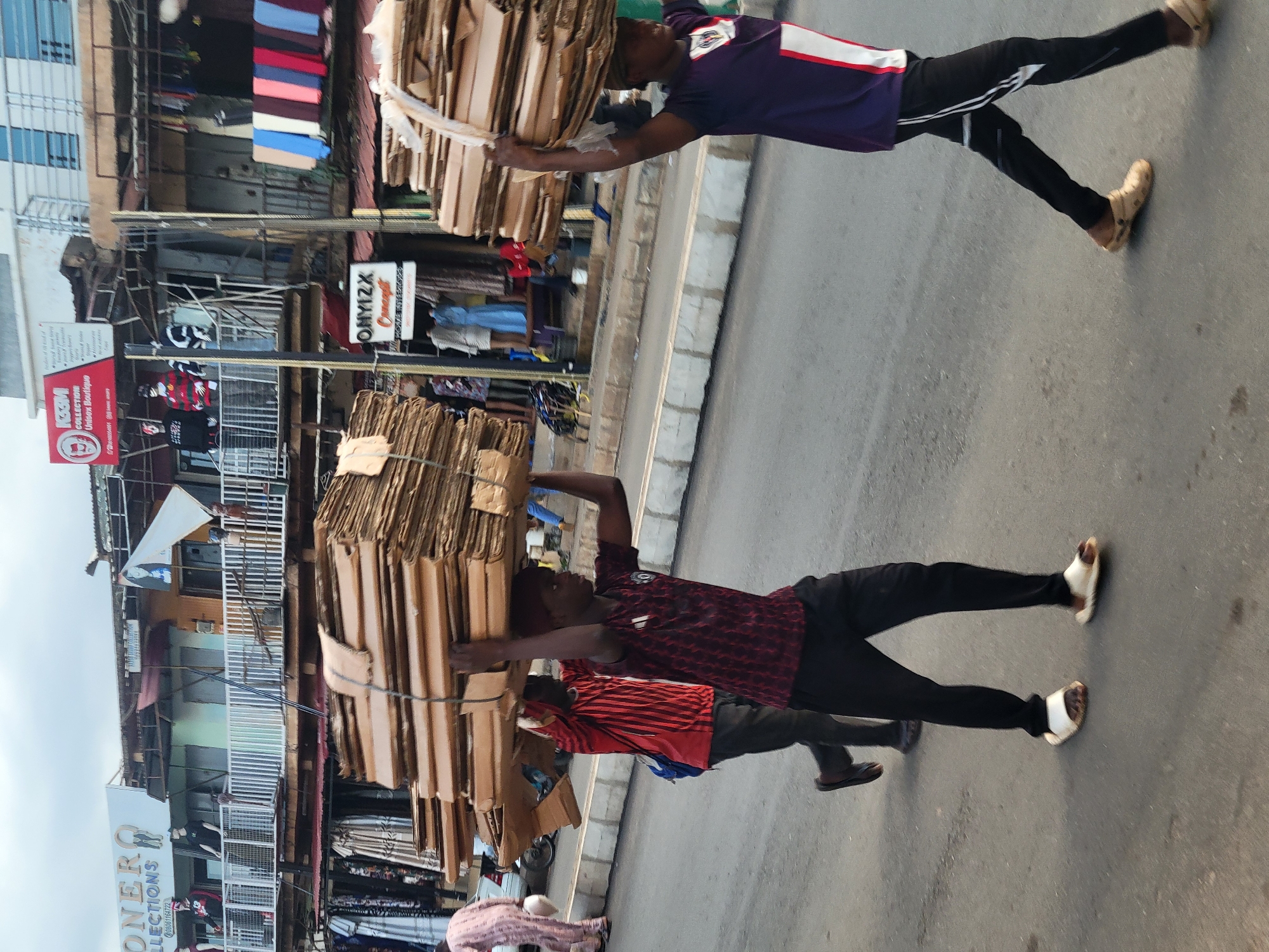 In Jos, Nigeria, two young men carry large stacks of cardboard on their heads along a city street. The man in the foreground, wearing a dark purple t-shirt with red and white accents and black pants, walks with a stack of cardboard tied with plastic wrap. Behind him, a second man in a red and black striped t-shirt and black pants also carries a similar load. A third person, partially visible in the background, walks away from the camera. The street is paved, and a raised concrete curb runs along the side. Buildings line the street, featuring shops with displayed clothing and signage, including "BONERO COLLECTIONS" and "K33M COLLECTION Unisex Boutique." The sky is overcast, suggesting a daytime scene.