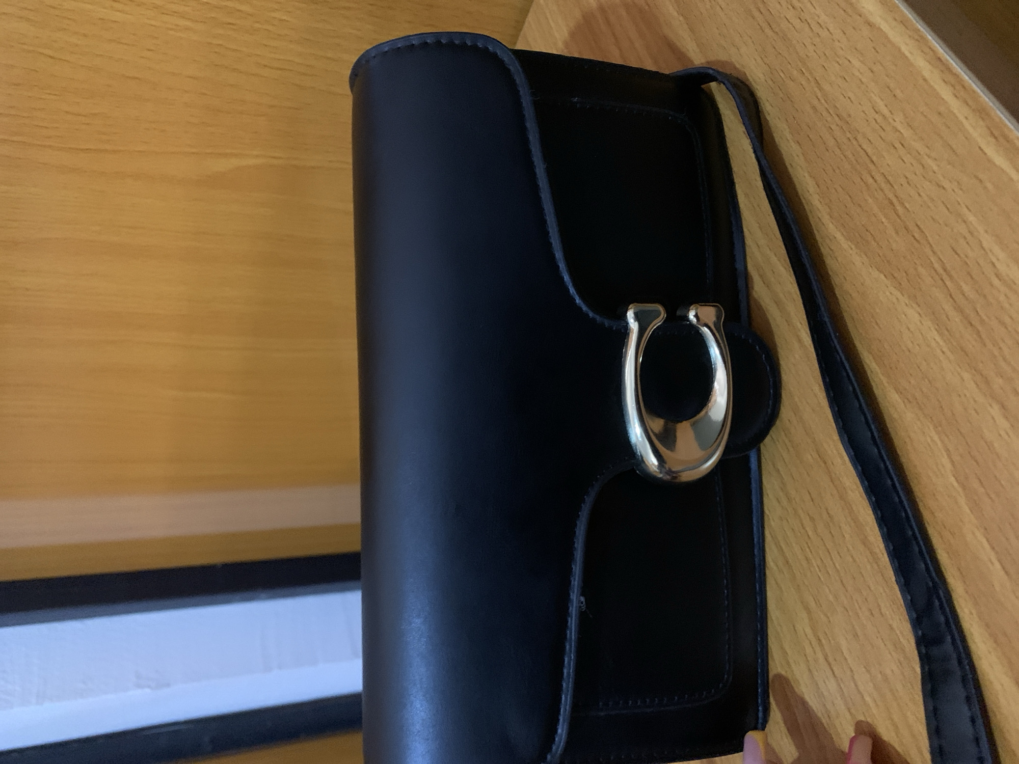 A black, rectangular, leather purse with a silver "C" logo clasp is placed on a wooden surface. A thin, black strap extends from the purse and lies diagonally across the surface. The background consists of vertical wooden planks. The lighting is warm and casts subtle shadows. The image appears to have been taken indoors.