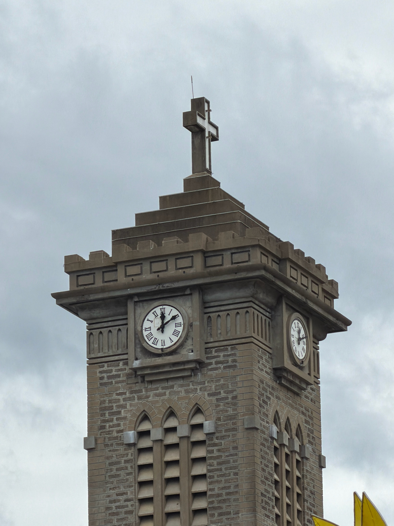 The image shows the upper section of a church tower, likely belonging to the Nha Trang Cathedral, in Nha Trang, Vietnam. The tower is constructed of brick and stone, featuring decorative battlements at the top. Two clock faces with Roman numerals are visible on the tower, with one clearly showing the time as approximately 11:58. A large white cross adorns the very top of the tower. The sky is overcast, with grey clouds dominating the background. The overall impression is one of a cloudy day, with a focus on the architectural details of the church tower.
