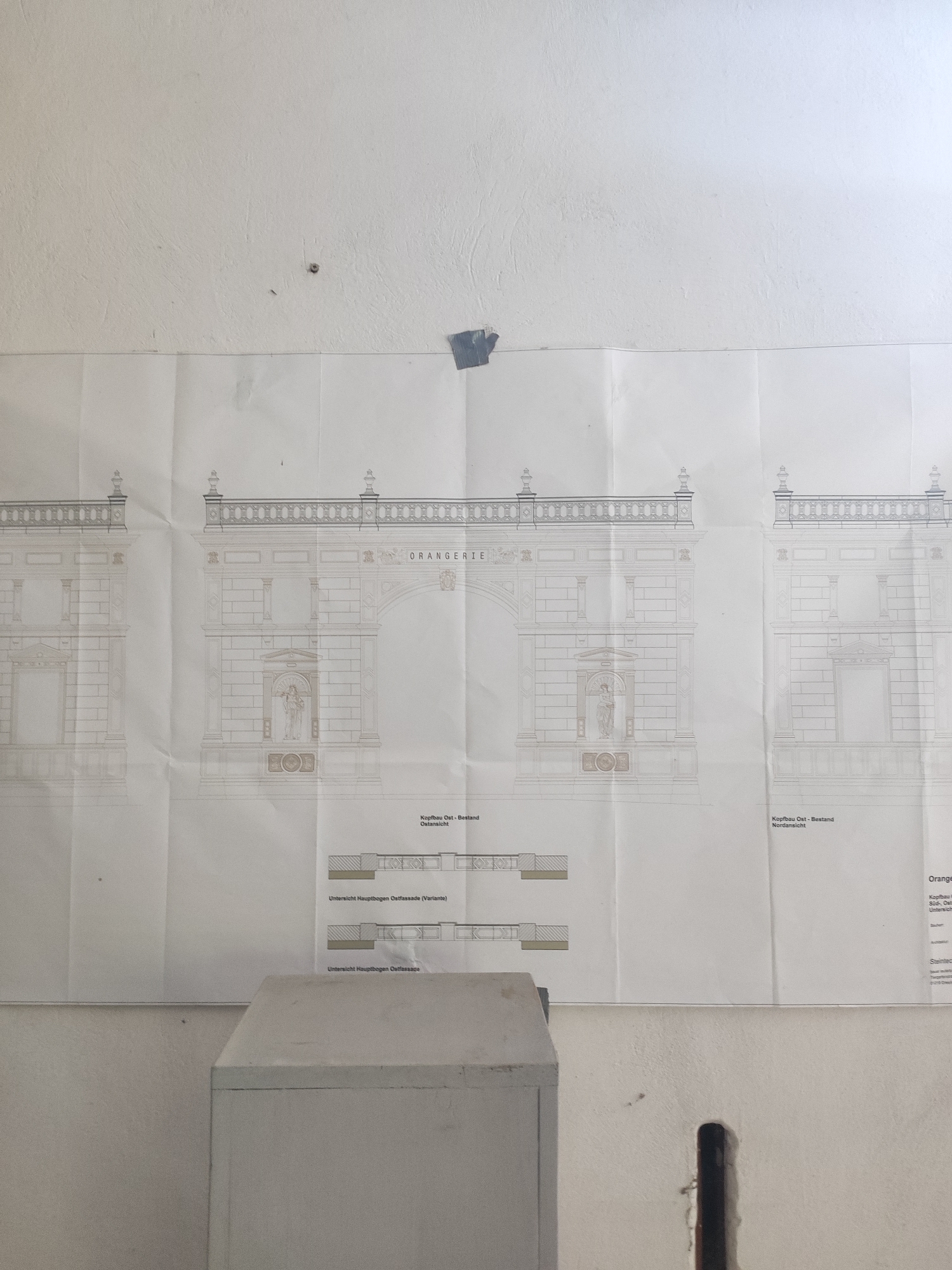 The image shows an architectural blueprint of the Orangery building at the Zwinger palace complex in Dresden, Germany. The blueprint depicts elevations and cross-sections of the structure, featuring classical architectural elements like columns, arches, and sculptures. The blueprint is taped to a textured, off-white wall, with a small, dark hole visible to the right of the blueprint. A light grey, box-like object is positioned in the foreground, obscuring the lower portion of the blueprint. The lighting suggests an indoor environment, possibly a workshop or storage area.