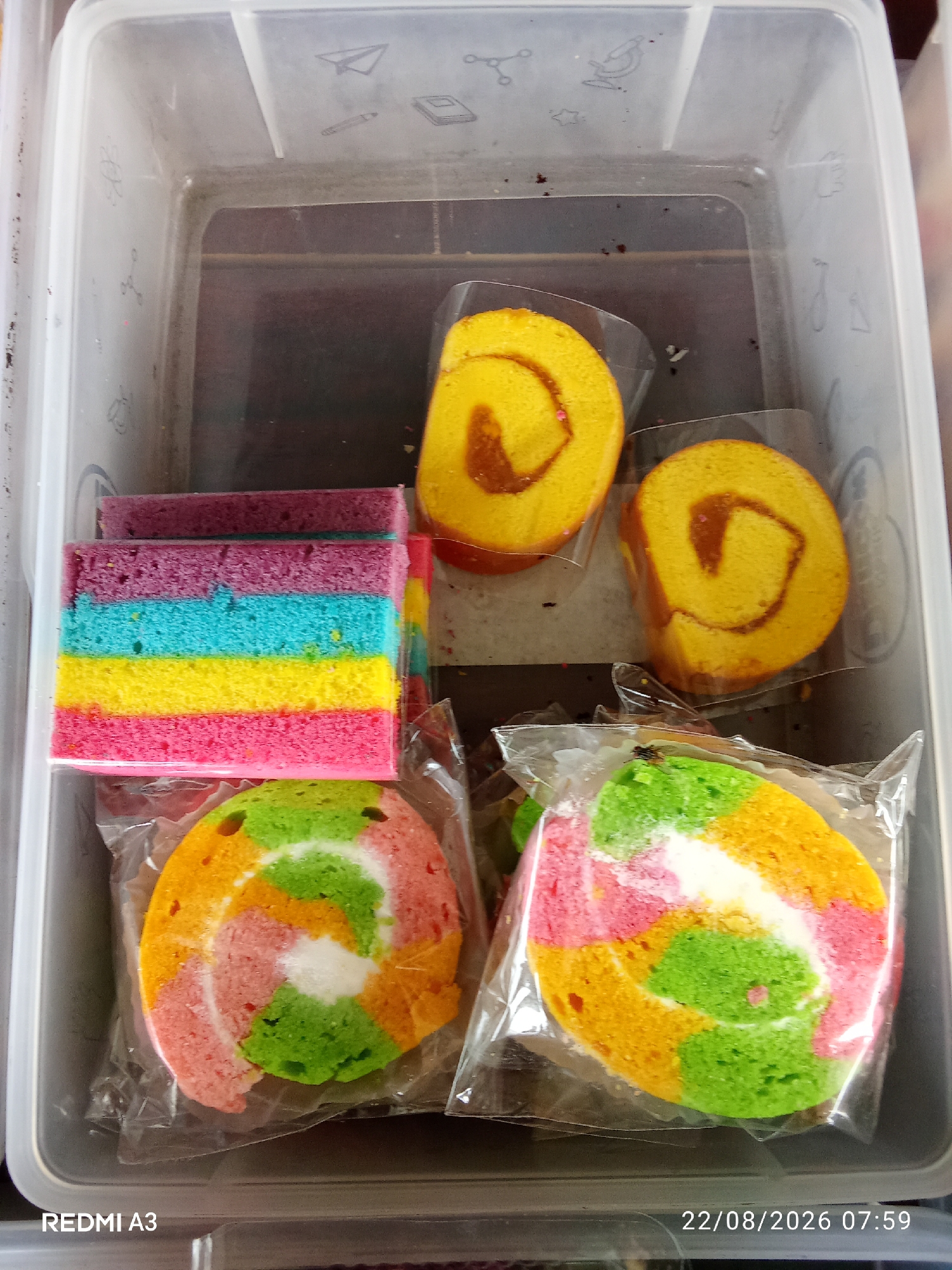 In a clear plastic storage container, a collection of colorful desserts is arranged. The container's lid features faint, embossed drawings of scientific and school-related items. In the upper portion of the container, two slices of yellow roll cake with an orange filling are visible, each individually wrapped in clear plastic. To the left of these, a stack of rectangular rainbow cake slices, featuring pink, yellow, blue, and purple layers, is also plastic-wrapped. The lower section of the container holds two circular rainbow roll cakes, each wrapped in clear plastic. These cakes exhibit a vibrant swirl of pink, yellow, green, and white colors. The background is a dark, blurred surface. The timestamp "22/08/2026 07:59" and the text "REDMI A3" are overlaid at the bottom of the image. The scene appears to be indoors, possibly in a home or a shop in Cemani, Indonesia.