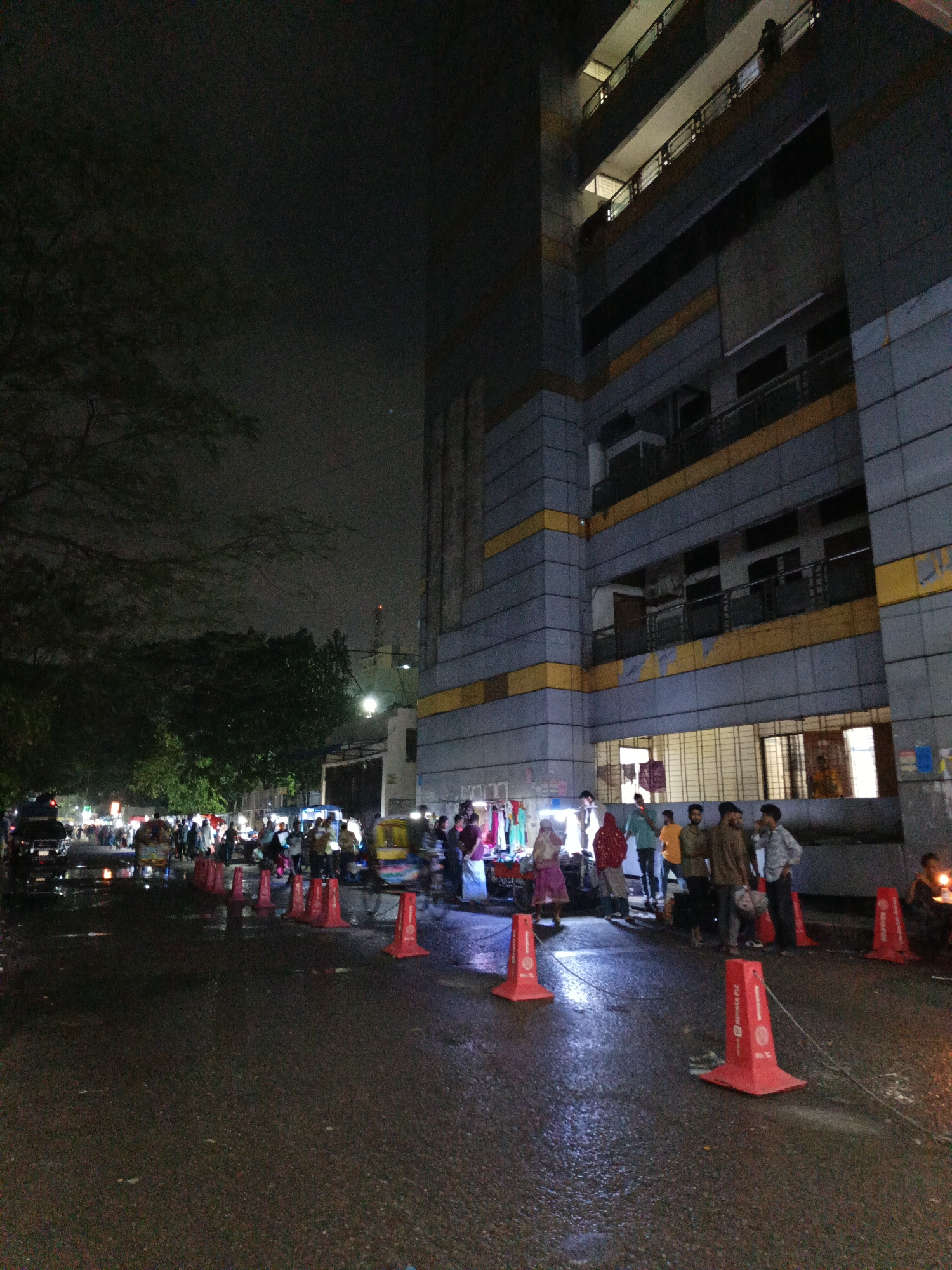 At night in an unknown city, a street market occupies a wet road alongside a tall, modern building with multiple stories and balconies. A row of orange traffic cones lines the road. People are gathered around market stalls, browsing or interacting with vendors. Some are standing and talking. The overall lighting is dim, with streetlights providing illumination. Trees line the opposite side of the road from the building.