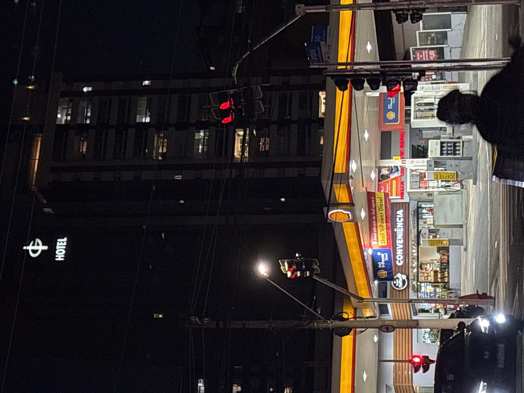 The image shows a nighttime scene at a Shell gas station in São Paulo, Brazil. A tall, modern building with lit windows is visible in the background, with a sign at the top that reads "HOTEL" and a symbol above it. In the foreground, the gas station features illuminated pumps and a convenience store. A traffic light is visible, showing a red light. A black car with its headlights on is stopped at the intersection. A person is silhouetted in the lower right corner, appearing to be seated. The overall atmosphere is urban and busy, despite the late hour.