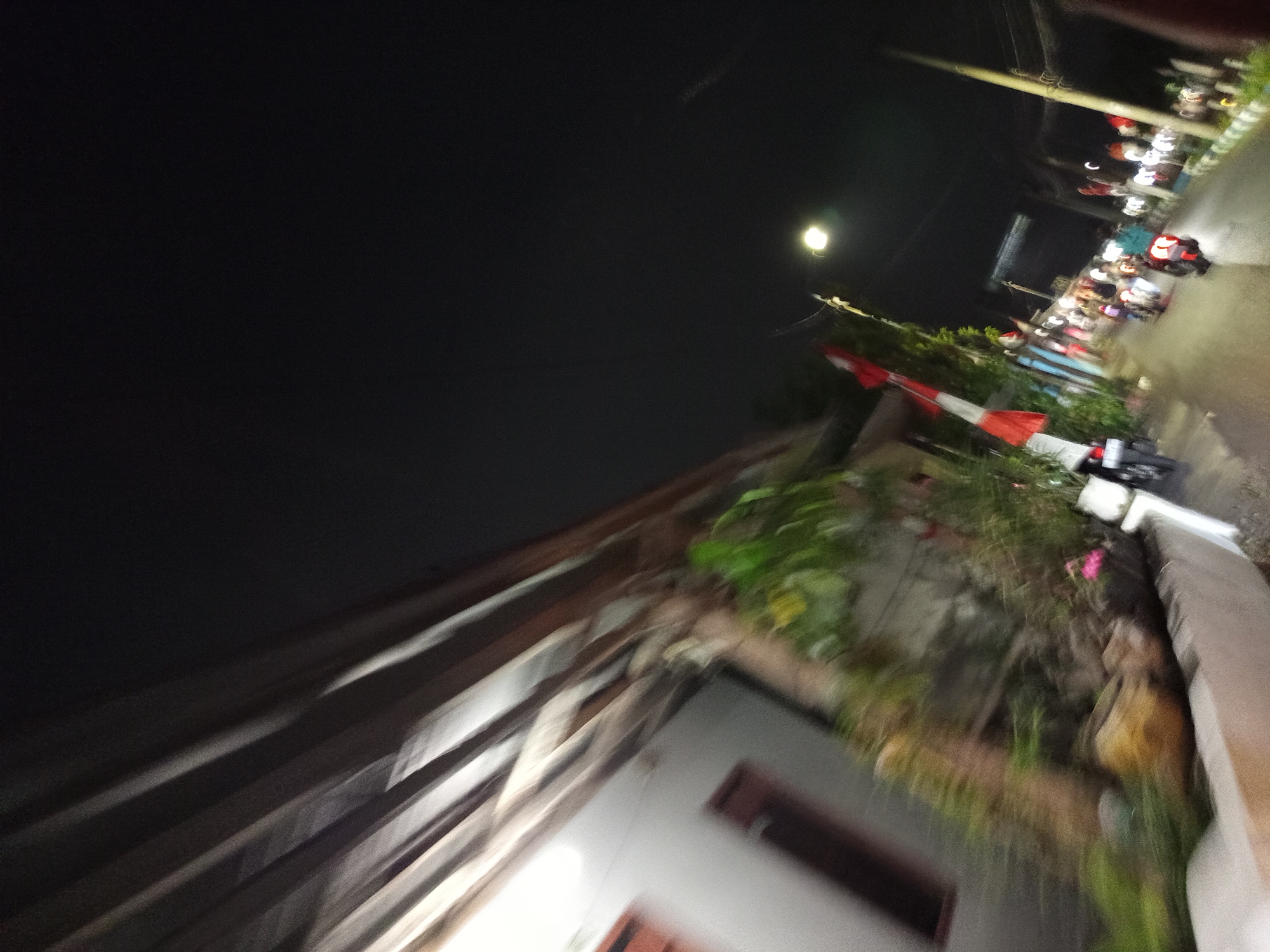 The image depicts a night scene on a street in Sruni, Indonesia, characterized by motion blur due to movement during capture. A building with a white wall and dark-framed windows is visible on the left. Vegetation, including trees and plants, lines the side of the road. Several motorcycles with riders are moving along the street, suggesting a procession or a busy commute. Decorative elements, including Indonesian flags (red and white), are present along the roadside. Streetlights and the lights from the motorcycles illuminate the scene against the dark night sky. The overall impression is of a lively, albeit slightly chaotic, nighttime atmosphere on a local Indonesian street.