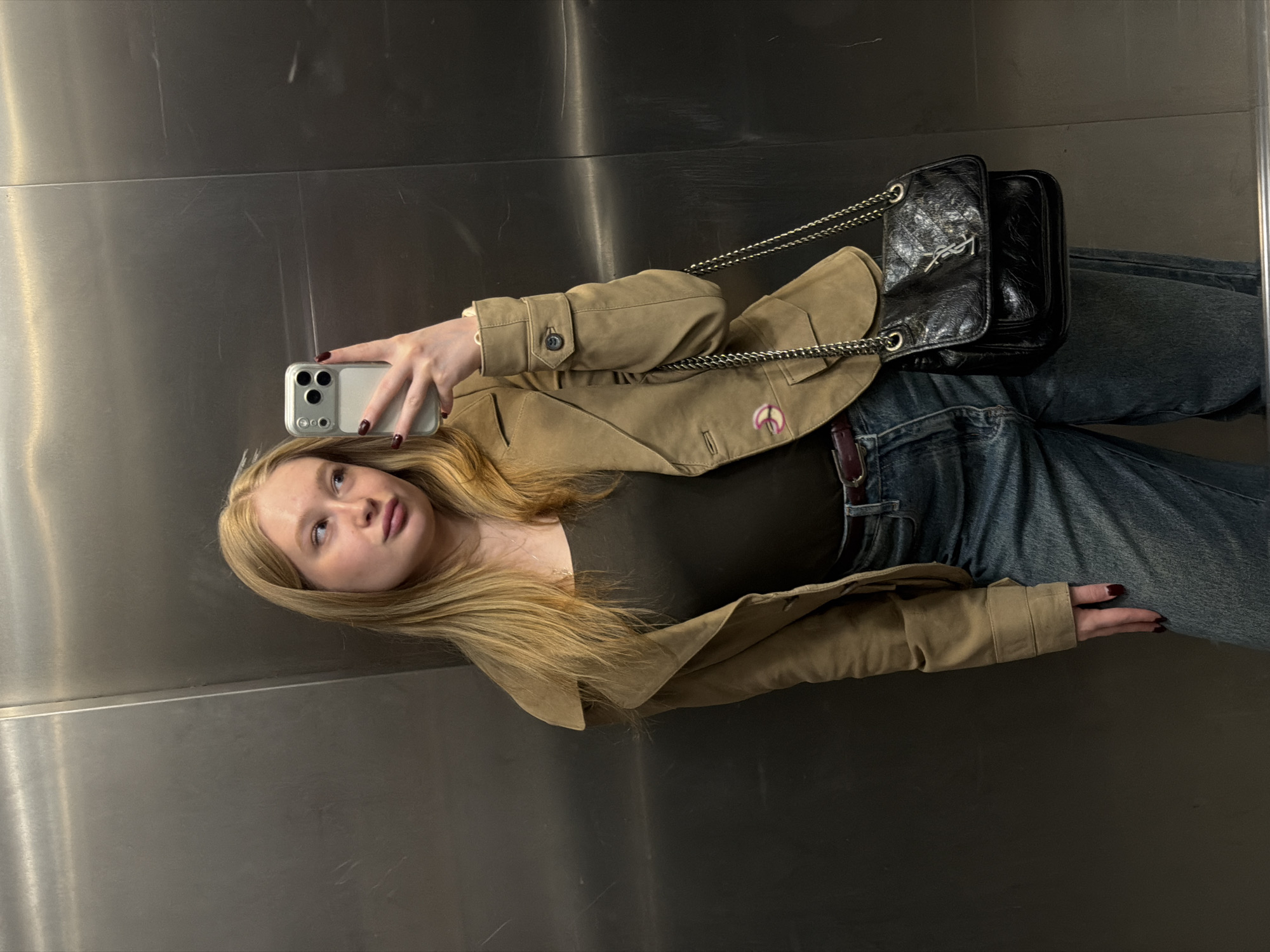 A blonde woman takes a selfie in an elevator with gray walls. She wears blue jeans, a brown t-shirt, and a brown jacket. A black handbag hangs off her shoulder. The location is not identifiable as being in Засечное, Russia.