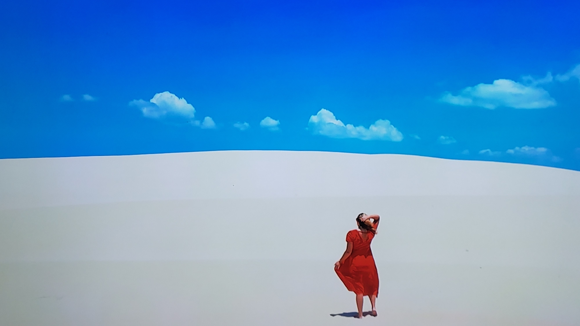A woman in a red dress stands on a white sand dune under a bright blue sky with scattered white clouds. The location is not Yerevan, Armenia.