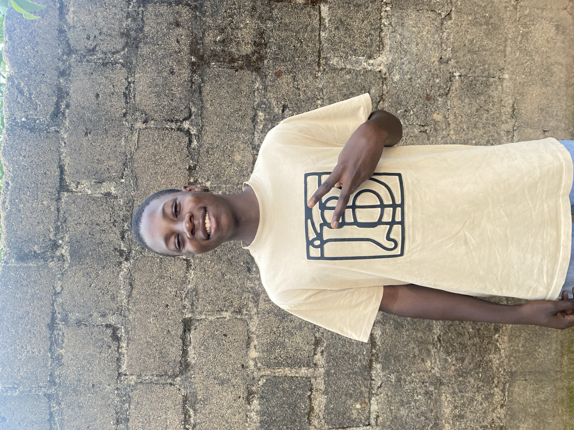 A young Black male stands in front of a rough, gray cinder block wall, likely outdoors. He is wearing a light tan t-shirt with a large, black graphic design on the front, possibly a stylized logo. He is smiling and holding up two fingers in a peace sign with his right hand, which is positioned over the graphic on his shirt. The lighting suggests it is daytime, and there is some greenery visible at the top edge of the frame, indicating an outdoor setting. The scene is situated in Gura Top, Nigeria.