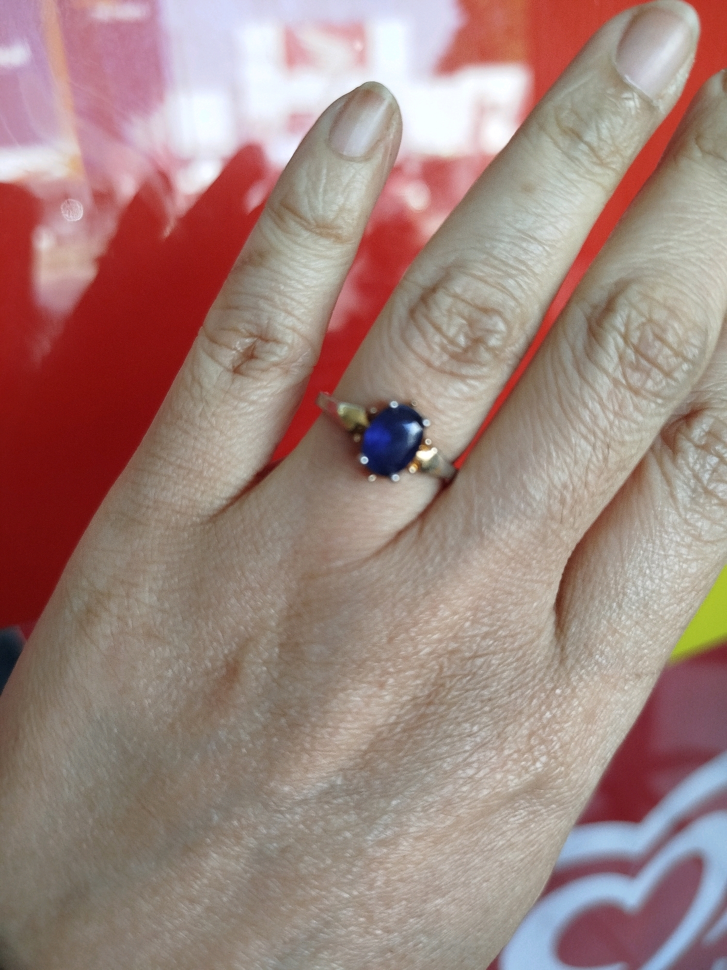 The image shows a close-up of a person's hand, adorned with a ring on the ring finger. The ring features a large, oval-shaped, deep blue gemstone, possibly a sapphire, set in a silver or white gold band with small, gold-colored accents on either side of the stone. The hand is resting against a bright red, reflective surface, which appears to be a car or a similar glossy object. The background is blurred, but hints of white and yellow suggest an outdoor setting, possibly in Sukomulyo, Indonesia. The lighting creates reflections on the red surface and highlights the texture of the skin.