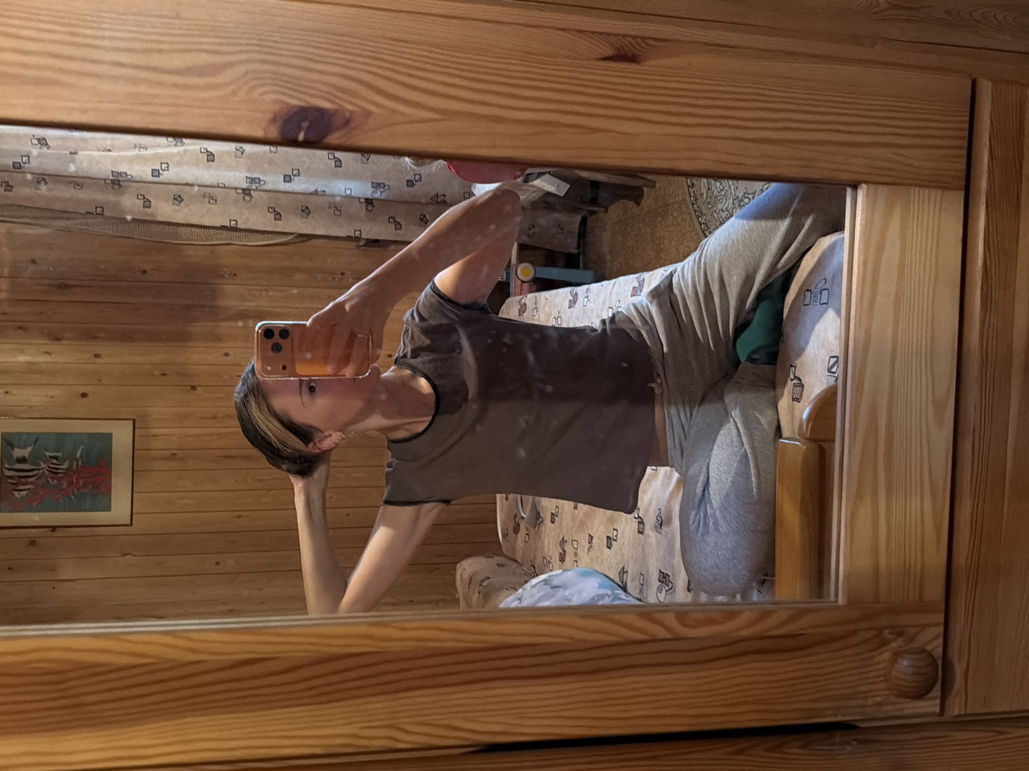 A woman is taking a mirror selfie in a bedroom. She has blonde hair pulled back and is wearing a gray t-shirt and gray sweatpants. She is holding a cell phone in her right hand and is using her left hand to adjust her hair. The room has wood-paneled walls and a bed with a patterned comforter. A framed picture of fish hangs on the wall. The mirror is framed in wood. The scene is indoors, likely in a residential setting. The city of Udelnaya, Russia is not discernible from the image itself, as there are no specific landmarks or identifying features.