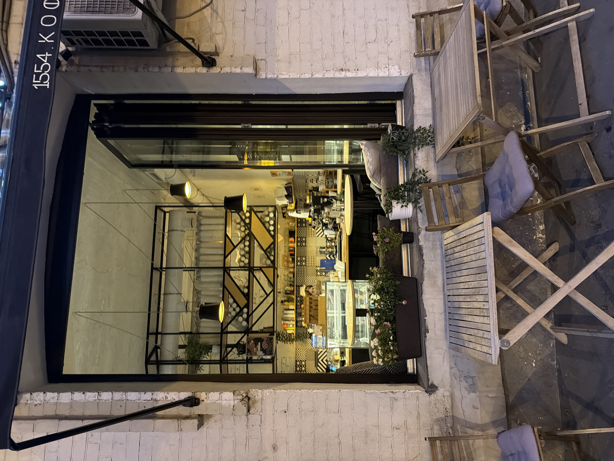 The image captures the entrance to "1554.KO," a cafe in Moscow, Russia. The cafe's storefront, marked with the name "1554.KO" on the awning, opens up with large glass doors, offering a view into the interior. The interior features a counter area and shelving stocked with various items. Several hanging lights illuminate the cafe's interior. Outside, the street-level area is furnished with wooden tables and chairs. The overall setting conveys an outdoor seating area of a cafe in an urban environment.