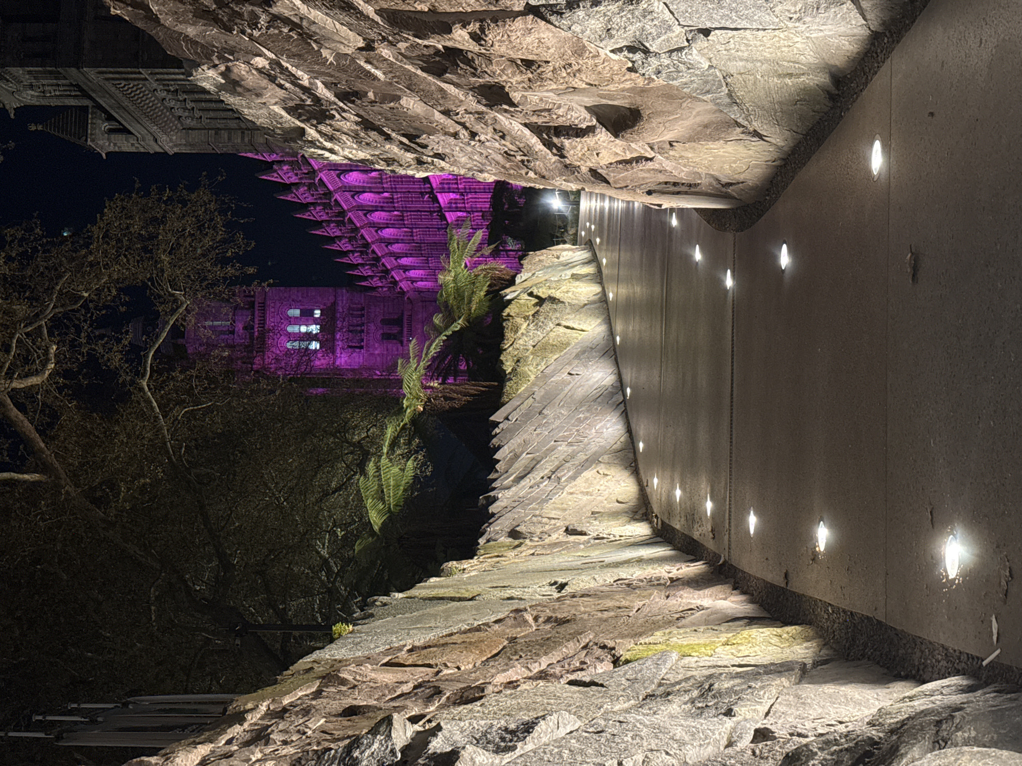 The image depicts an outdoor scene at night in London, United Kingdom. A stone walkway, illuminated by recessed lights along a concrete wall, leads towards a building. The building is bathed in purple light, likely the Tower of London. Trees are visible to the left, silhouetted against the night sky. The perspective is unusual, as the image appears to be rotated. Stone walls flank the walkway. No people are visible.