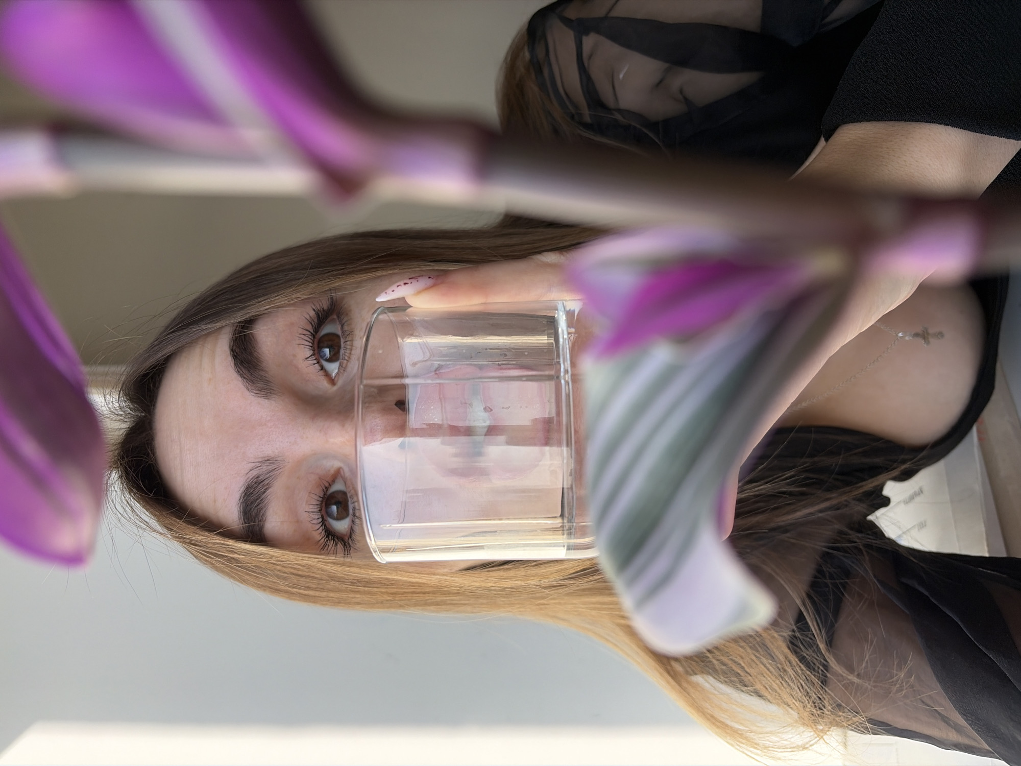 A woman is holding a glass of water in front of her face. She has brown eyes, and light brown hair. She is wearing a black top and a silver necklace with a cross pendant. Her fingernails are long and painted. There are purple flowers in the foreground, slightly out of focus. The background is a blurred-out, light color. There is no discernible location information or details related to Izhevsk, Russia.