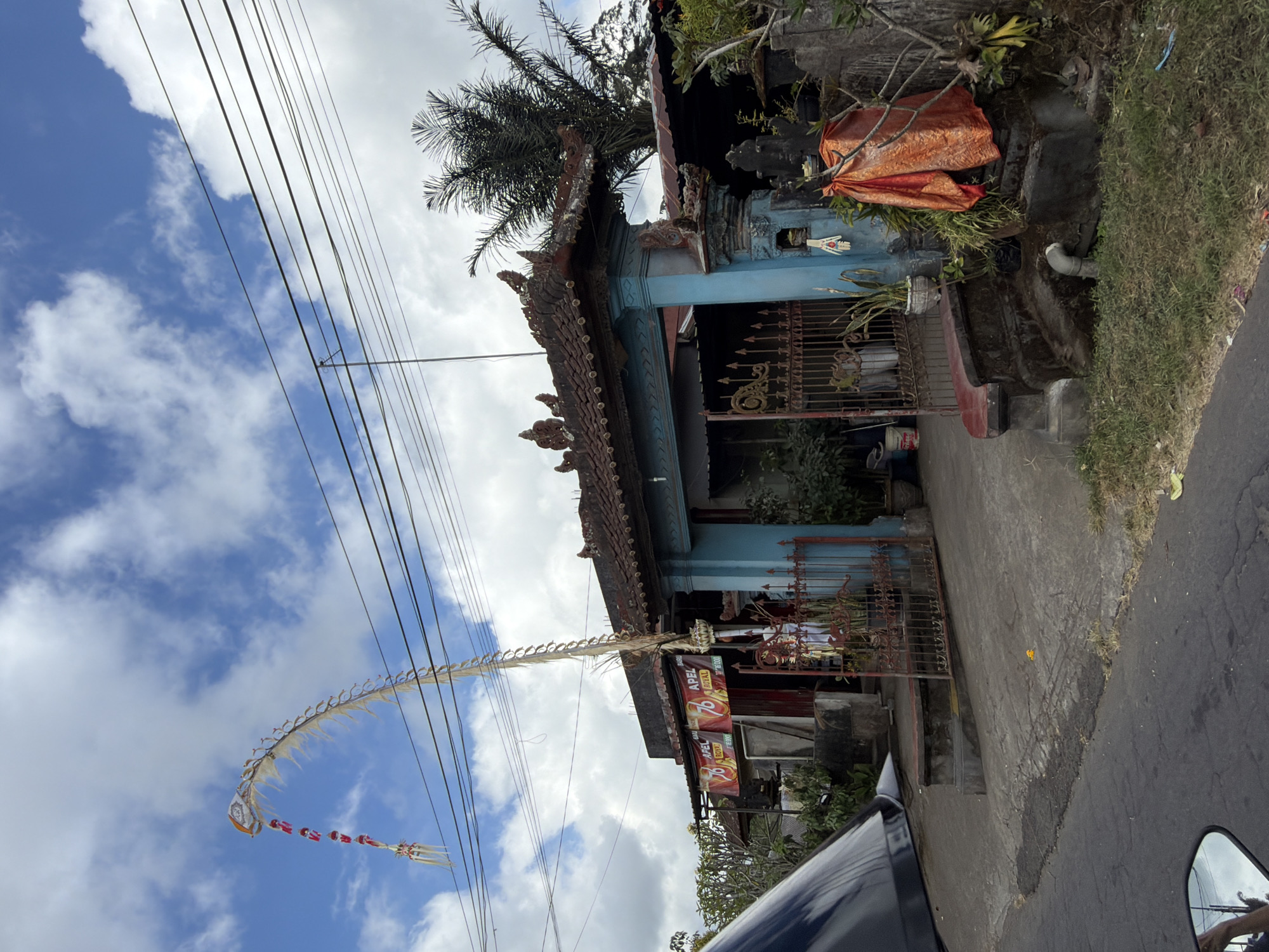 The image depicts a street scene in Kintamani, Indonesia, featuring a traditional Balinese gate and storefront. Dominating the sky is a large, ornate kite, likely a Penjor, a ceremonial bamboo pole decorated with offerings, which arcs gracefully across the frame. Below the kite, a building with a red-tiled roof and light blue decorative columns serves as the entrance. A red metal gate is partially open, revealing a glimpse of the interior, which appears to be a shop or residence with plants and various objects. Two banners with the number "76" and text reading "APEL BARU ROYAL" are visible on the storefront. To the right of the gate, a statue draped in an orange cloth stands near some overgrown vegetation and a concrete drainage pipe. A palm tree is visible behind the building. The street in the foreground is paved with asphalt and shows some signs of wear, with grass growing along the edges. The overall impression is of a typical, culturally rich streetscape in a rural or semi-rural Indonesian setting.