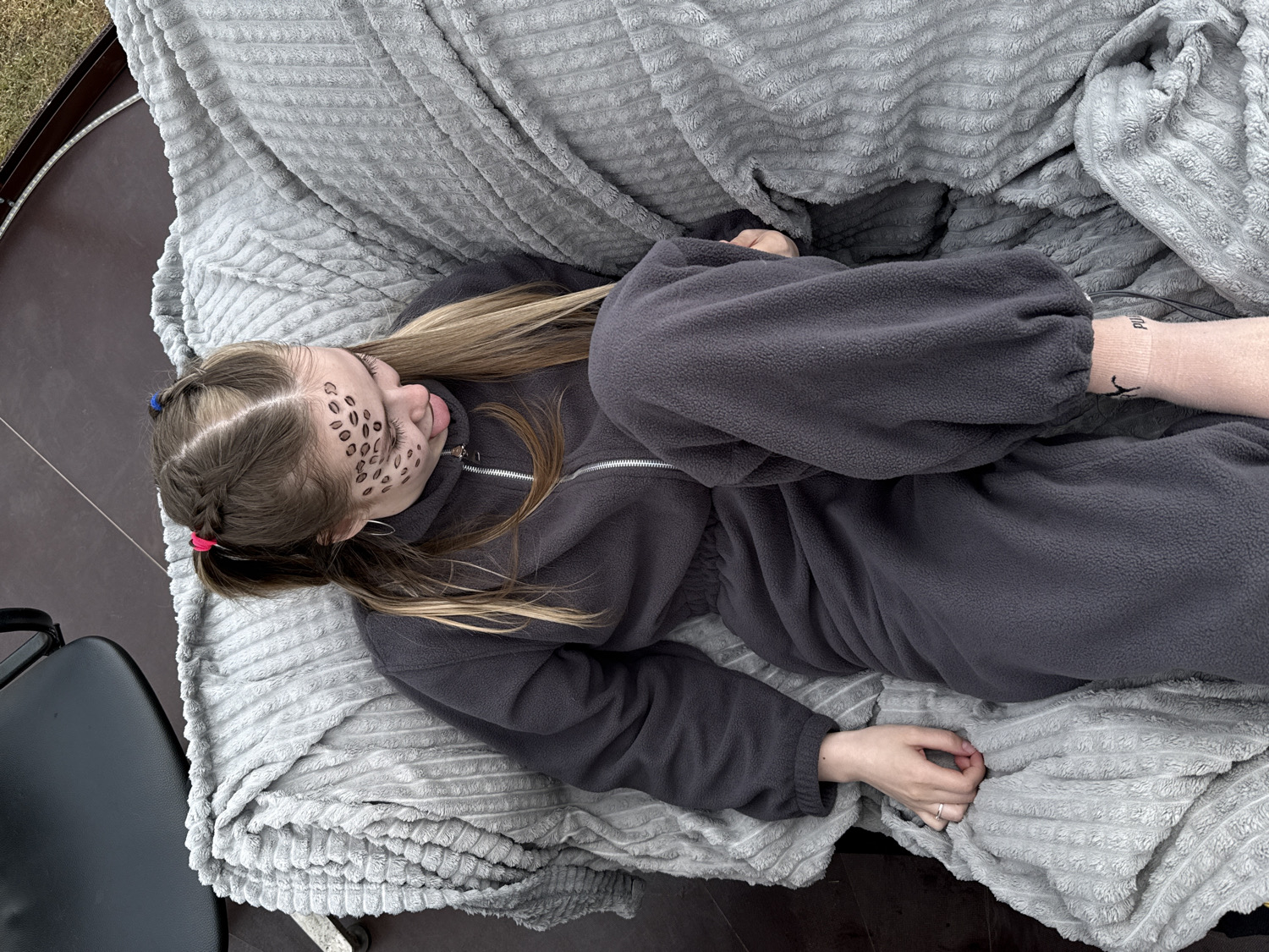 The image displays a young individual lying comfortably on a soft, grey textured blanket, likely resting or relaxing in an outdoor or semi-outdoor setting in Zhigalki, Russia. The person is dressed in a dark grey fleece jumpsuit or loungewear, conveying a sense of warmth and comfort.
The individual's blonde hair is partially pulled back into two small braids, accented with blue and pink hair ties, while the rest cascades loosely. A notable detail is the playful face paint on their right cheek, featuring multiple small, elongated dark markings resembling coffee beans or leopard spots. Their tongue is slightly extended, giving a relaxed or whimsical expression. One hand, adorned with a ring, rests on the blanket, and a light-colored sock with the partially visible brand name "Puma" printed on it is seen on one foot.
The setting appears to be illuminated by natural daylight, suggesting it is daytime. The blanket is spread over a dark brown surface, possibly an outdoor table or wooden deck, with a glimpse of green grass visible in the upper left background. A portion of a black chair or stool is also present on the left. The scene evokes a casual and relaxed atmosphere, possibly during leisure time or a playful event.