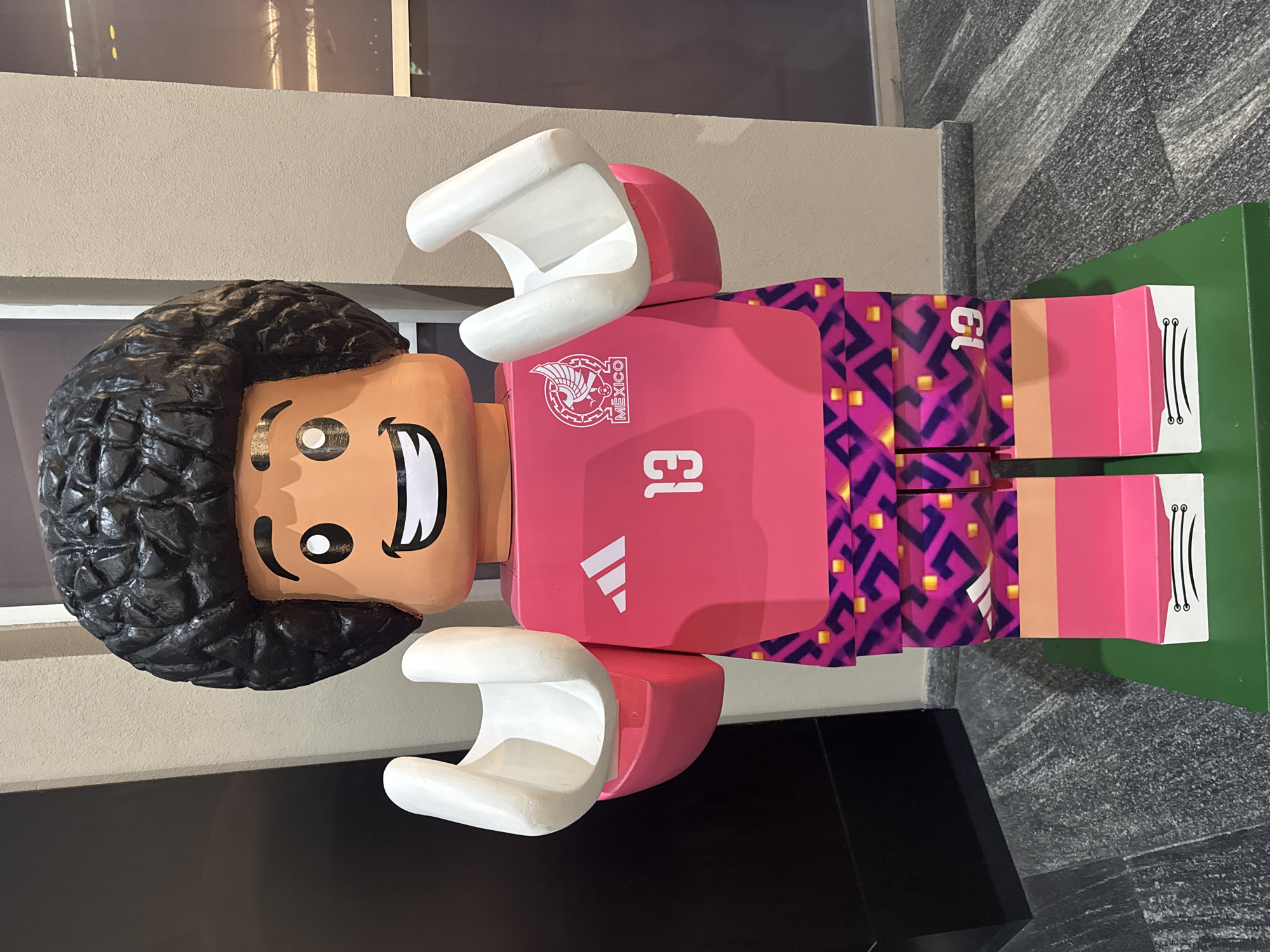 A large, painted Lego figure of Guillermo "Memo" Ochoa, the goalkeeper for the Mexican national soccer team, is presented indoors. The Lego figure is wearing the pink goalkeeper jersey with the number 13 and the Mexican soccer federation logo. The background consists of a concrete wall and a granite tile floor. The location is Puebla, Mexico, evident from the team logo. The figure is mounted on a green base.