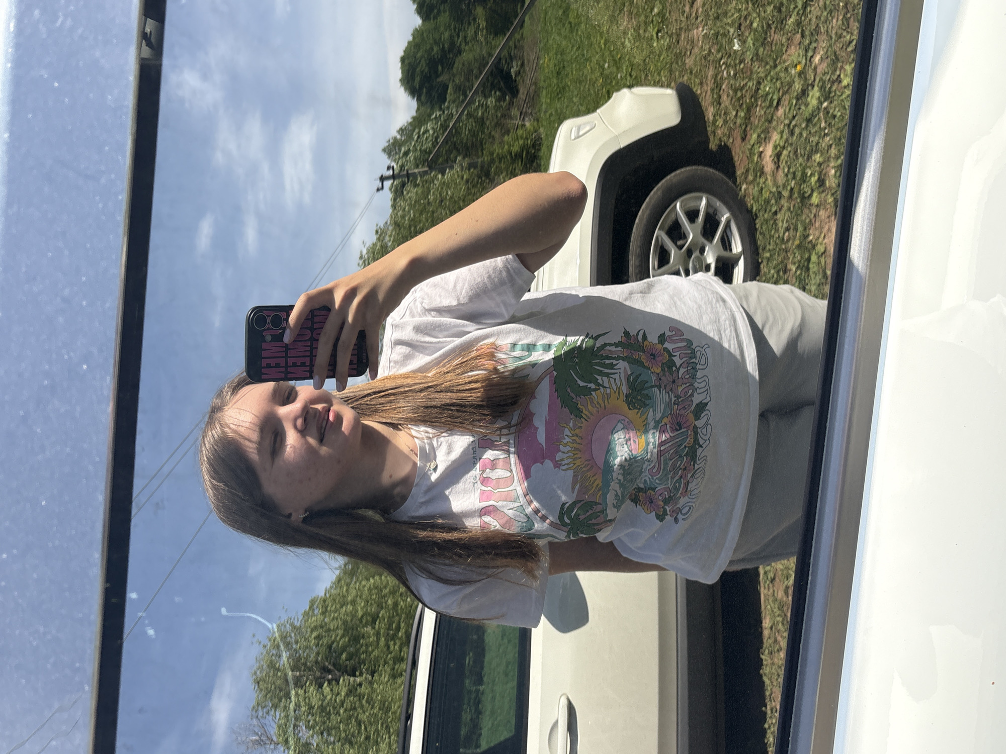 A young person is taking a selfie in a mirror. They are wearing a white t-shirt with a colorful graphic print. Their hair is light brown and long. They are holding a smartphone with a black case. The reflection in the mirror shows a white vehicle, likely a SUV or crossover, parked in a grassy area with trees in the background. The sky is visible with some clouds. The photograph appears to be taken outdoors. The location is not discernible from the image.