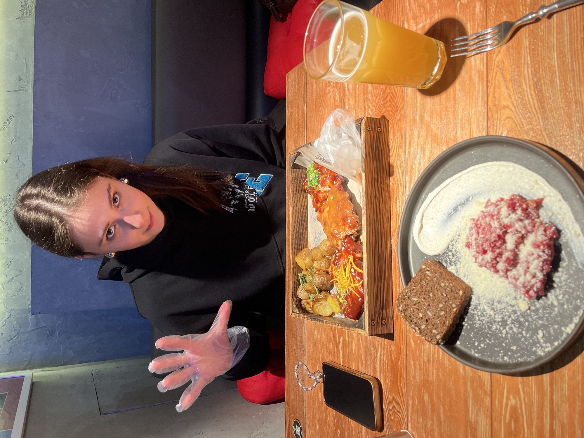 The image depicts a female individual seated at a wooden table in an indoor restaurant setting in Родниковское сельское поселение, Ukraine. The subject, a young woman with long dark hair, is positioned centrally and looks directly at the camera. She wears a black hoodie with a light blue and white graphic. Her right hand, wearing a clear disposable plastic glove, is raised and open, facing the camera.
On the wooden table in front of her, various food items and personal belongings are arranged. A rectangular wooden tray holds roasted baby potatoes and two pieces of meat, possibly ribs, glazed with different sauces and garnished. One piece has yellow string-like garnish, and the other has green chopped garnish. Adjacent to the tray, a dark gray plate contains a portion of minced red meat, likely steak tartare, topped with grated white cheese, a dollop of white cream or sauce, and a slice of dark seeded bread. A tall glass filled with an amber beverage, consistent with beer, sits to the right of the food. A silver fork is placed nearby. A dark smartphone with a gold-colored frame and a sparkling ring or bracelet on top lies horizontally on the table to the left.
The background features a textured wall, split horizontally into an upper section of muted blue-green and a lower section of darker blue-gray. A red cushioned booth seat is visible behind the table. The lighting is dim, with prominent illumination on the table and the subject.