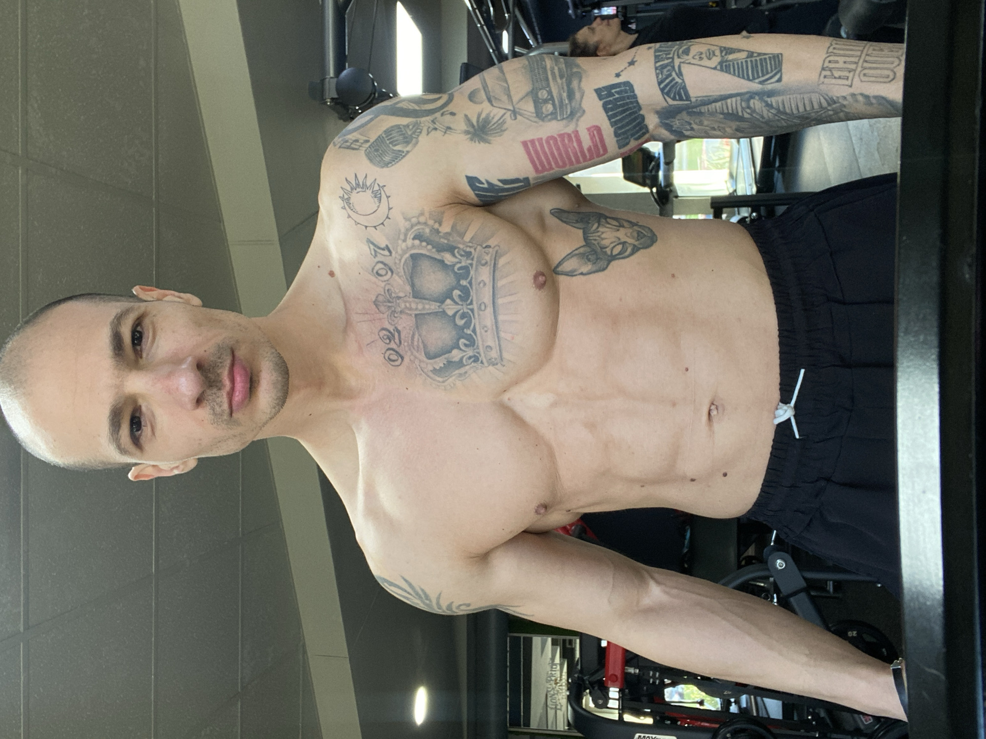 A shirtless man with a shaved head stands indoors. He has numerous tattoos on his arms and chest, including a crown on his chest, the word "WORLD" on his right arm, and other various designs. He is wearing black athletic shorts with a white drawstring. The man appears to be in a gym setting, with exercise equipment visible in the background. The location is not identifiable as Pleven, Bulgaria from the image alone.