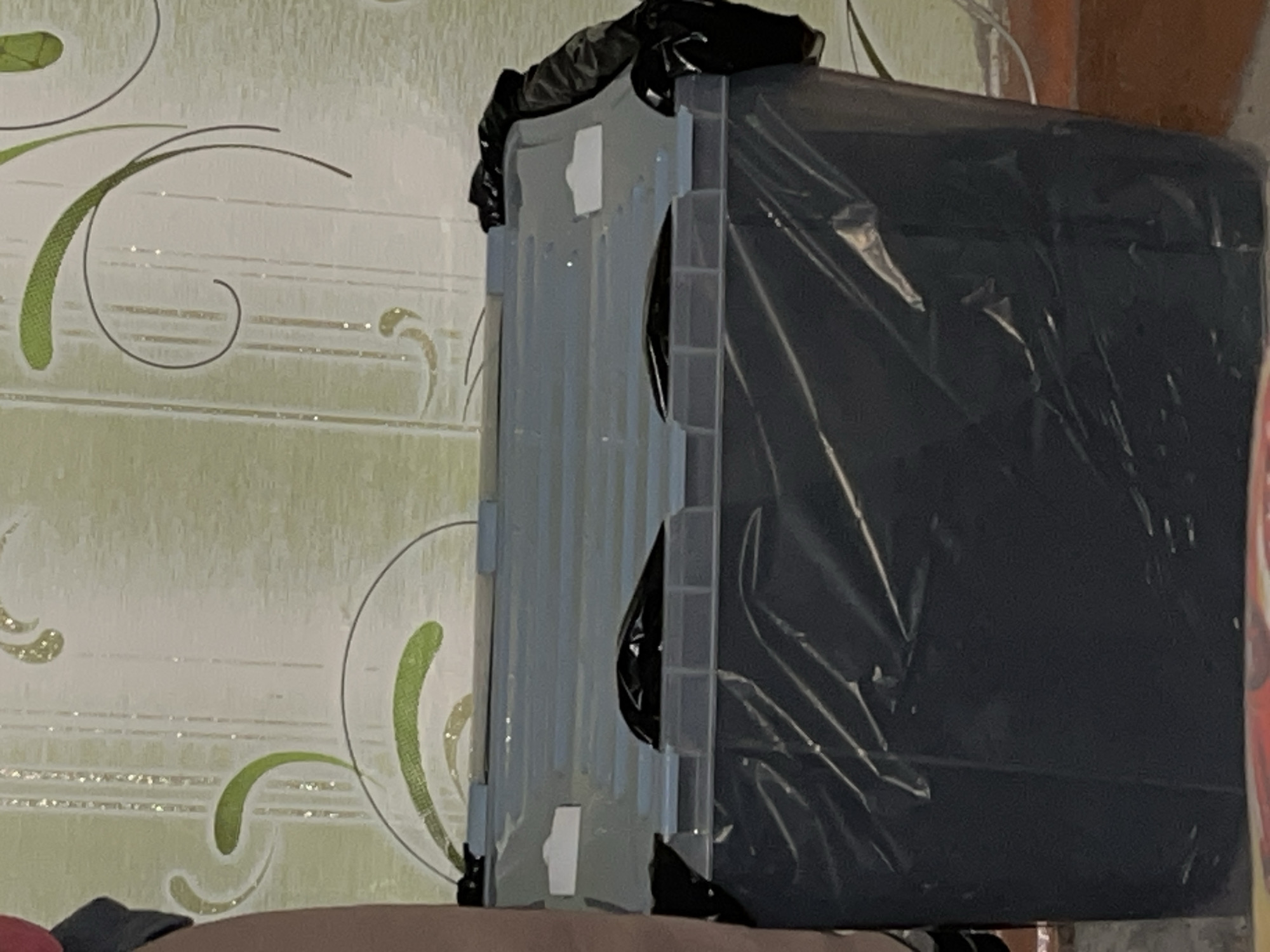 The image displays an indoor scene, likely in Minsk, Belarus, featuring a large, dark object on the right, completely enveloped in crinkled black plastic sheeting. This object, possibly a television or monitor, rests on a light-colored, flat surface, which could be part of a furniture item or another box.
To the left of the covered object, a light grey or translucent plastic storage container or crate is visible. It appears partially open or folded, with some dark material, possibly another black bag, draped over its top edge. The background consists of a wall covered in light-toned wallpaper. This wallpaper features a decorative pattern of flowing, stylized green and dark grey or black lines, resembling vines or swirls, interspersed with subtle glittery accents.
The lighting in the image is somewhat dim, suggesting either low ambient light or artificial illumination. No human subjects are present, and no discernible activity is taking place; the scene depicts a static arrangement of items. The plastic covering on the large object implies it is either being stored, protected from dust or damage, or prepared for transport. No visible text can be identified in the image.