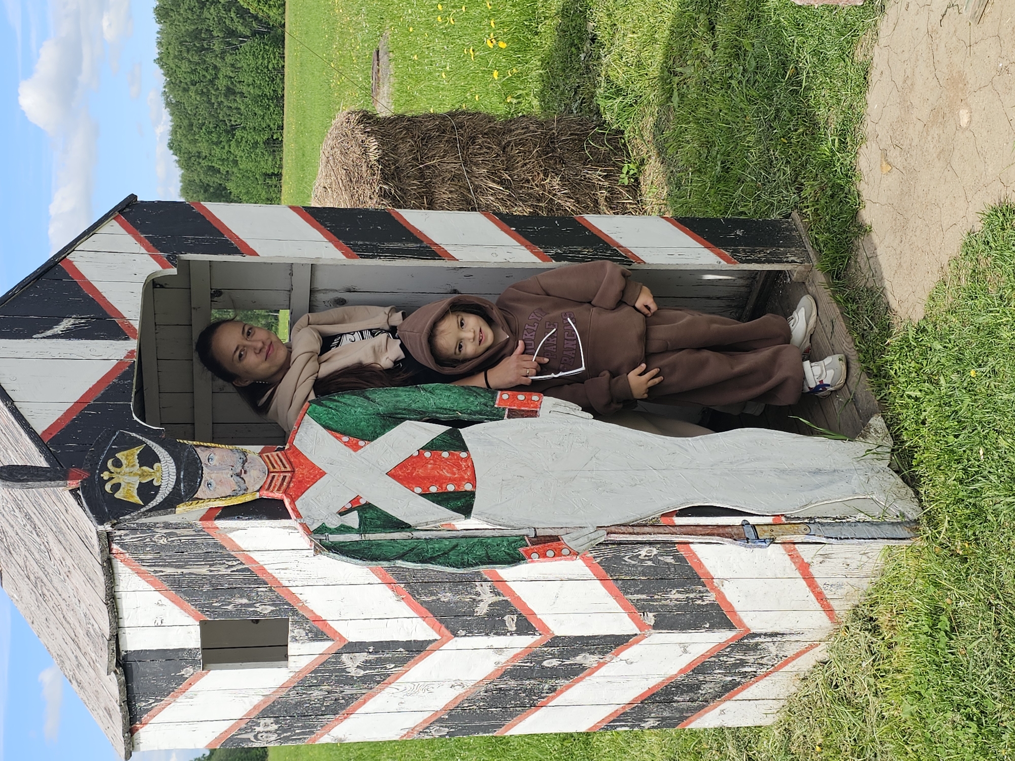 Two individuals, an adult female and a young child, are positioned within a wooden structure resembling a guardhouse. The structure is painted with black and white chevrons interspersed with red stripes. On the side of the structure, a painted depiction of a historical soldier is visible. The adult is lying behind the child who is in the foreground, each looking toward the camera. The environment includes a field with grass and a haystack visible in the background. The photograph was taken in the Borodinskogo Muzeya, Russia.