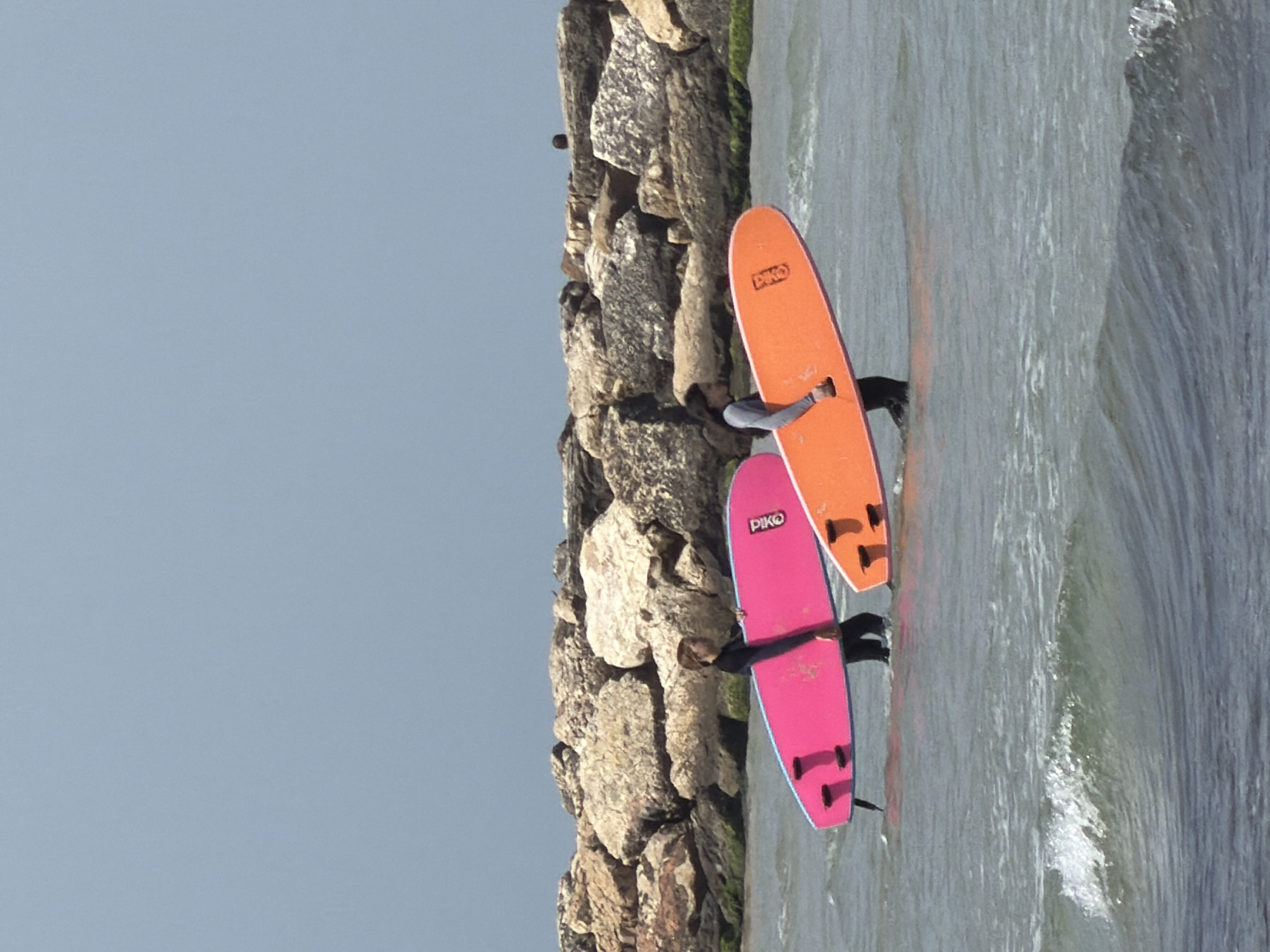The image depicts two individuals standing in shallow water with surfboards near a rocky breakwater. The location is likely a beach in Tel Aviv, Israel. One surfboard is orange and the other is pink, both bearing the word "Piko". The individuals are wearing wetsuits. The water is slightly choppy with small waves breaking. The sky is overcast. The rocky breakwater is covered with some green algae or moss.