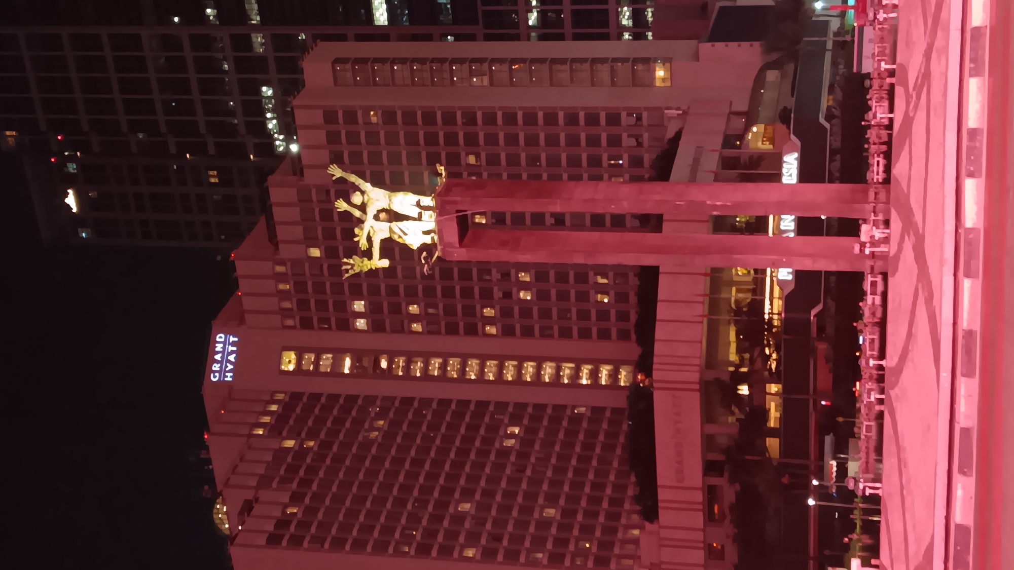 The image captures a night view of the National Monument (Monas) area in Central Jakarta, Indonesia. In the foreground, a pinkish glow illuminates a plaza area with what appears to be decorative pavement and some seating structures. Dominating the center of the frame is the iconic National Monument, a towering obelisk topped with a flame, rendered in white and illuminated from below. The monument stands on a red, rectangular pedestal, with a sculpture of three figures at its summit. Behind the monument, the Grand Hyatt hotel is prominently visible, its facade lit with warm yellow lights from numerous windows. The Grand Hyatt sign is clearly visible in white lettering against the pinkish hue of the building's exterior. To the right of the Grand Hyatt, another tall building with a grid-like pattern of windows is partially visible, also lit from within. The sky is dark, indicating it is nighttime. The overall scene is one of urban illumination and monumental architecture.