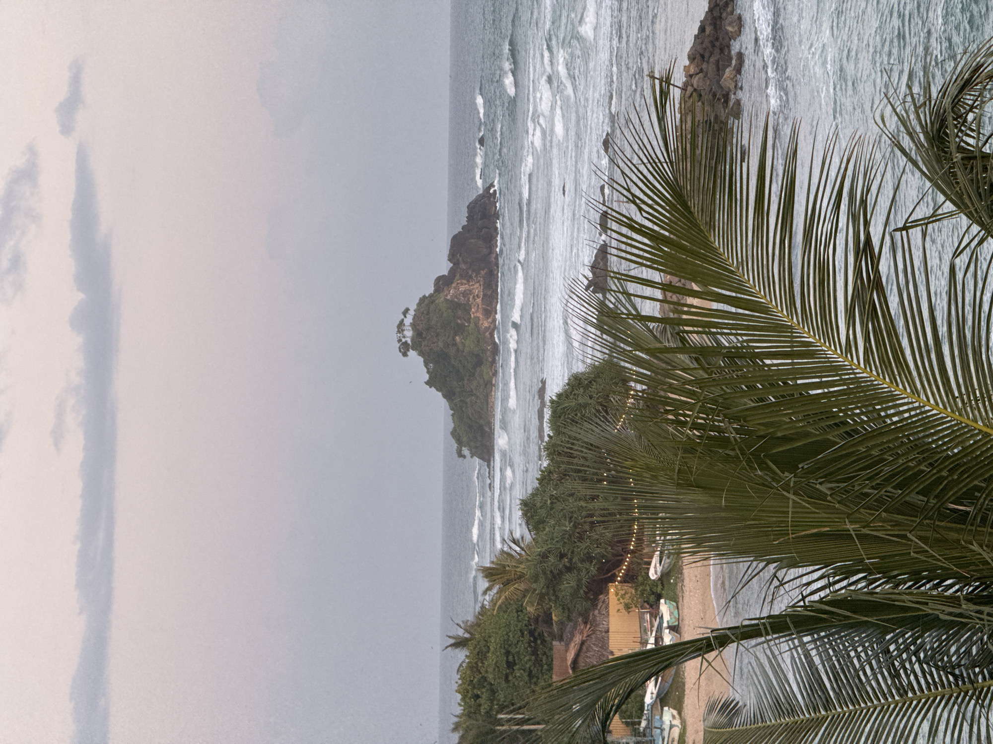 The image depicts a coastal scene in Goviyapana, Sri Lanka, at dusk. The sky is overcast with soft pastel hues. In the foreground, palm fronds obscure a portion of the view. Below the fronds, a beach is visible with several small, empty boats resting on the sand. Some of these boats are white with green trim. A thatched-roof structure and a fence line the edge of the beach, with string lights illuminating the area. Lush green foliage, including palm trees and dense bushes, separates the beach from the ocean.
The ocean itself is rough, with white-capped waves rolling towards the shore and around a rocky outcrop in the mid-ground. A small, densely vegetated island with a few trees on its summit sits offshore, further out in the sea. The water appears a deep blue-grey. The overall impression is one of a tranquil, albeit slightly moody, tropical evening.